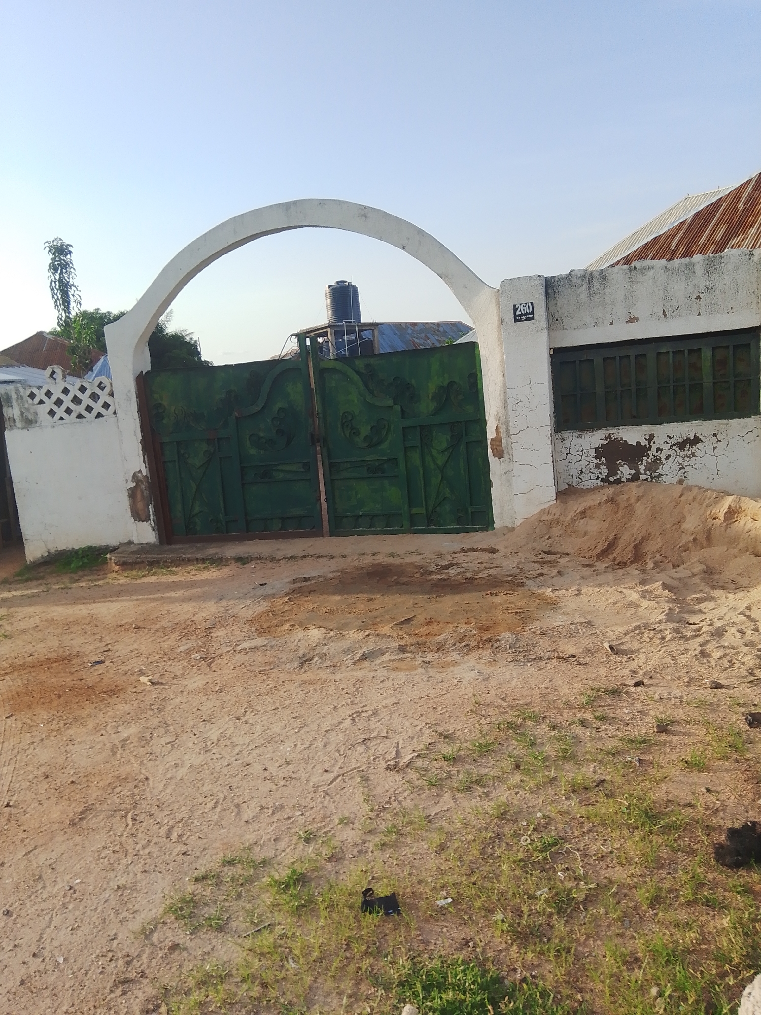The image shows a large, green metal gate with intricate scrollwork, framed by an arched white stucco archway. To the left of the gate, a white stucco wall with a diamond pattern adorns its top edge. A tall, slender tree with sparse foliage is visible behind this wall. To the right of the gate, another white stucco wall is present, featuring a dark rectangular sign with the number "260" in white lettering. Adjacent to this wall is a window with dark green bars. A pile of light brown sand sits in front of the right wall, and the ground in front of the gate is a dirt and sand mixture with patches of sparse green grass. A black water tank is visible on the roof of a building behind the gate. The overall scene depicts an entrance to a property, likely in a residential or commercial area, with a clear sky overhead.