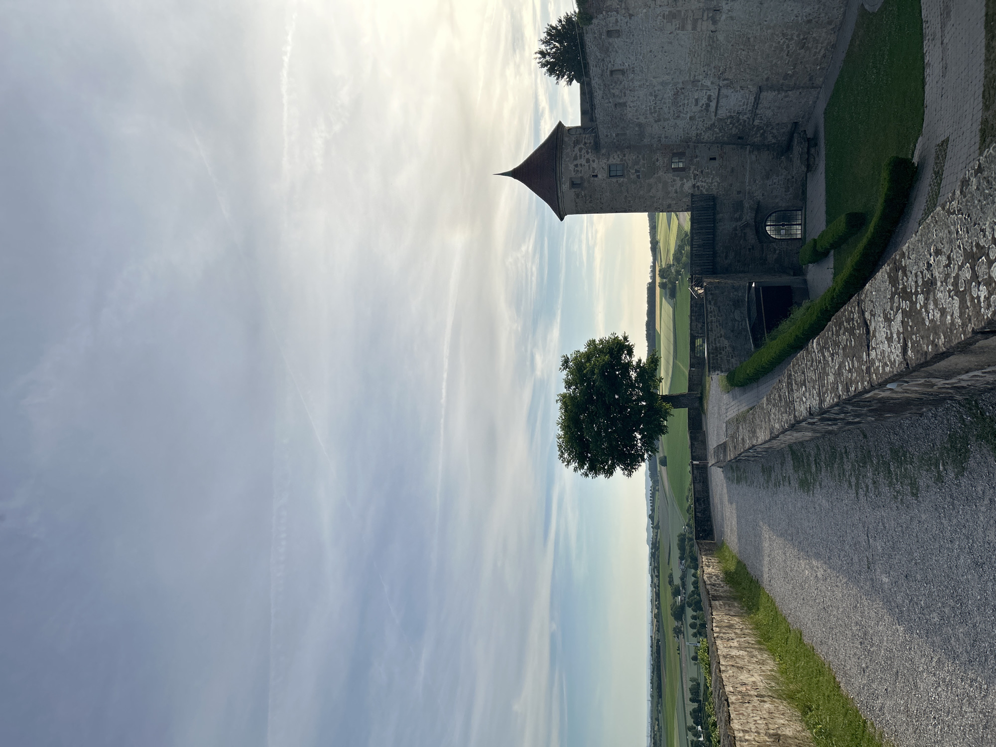 This image depicts an elevated view from the grounds of Stettenfels Castle in Untergruppenbach, Germany, overlooking a rural landscape under a partially clouded sky. The sky, occupying the upper two-thirds, transitions from pale blue-grey on the left to a bright, hazy white-yellow on the right where the sun is located, featuring wispy cirrus clouds and faint contrails.
In the foreground, a light grey gravel path, approximately 2.5 meters wide, curves to the left and recedes into the distance. It is bordered on the left by a stone wall, roughly 1 meter high, with grass growing along its base. On the right, a lower stone wall, about 0.5 meters high, shows visible lichen and moss growth on its top surface. Beyond this right wall, a manicured green lawn is partially visible, bordered by a dark green, sculpted hedge.
In the midground, on the right side, a cylindrical stone castle tower with a steep, conical reddish-brown tiled roof stands prominently. The tower features several small, rectangular windows. Adjacent to the tower is a larger section of the castle's stone wall, which includes an arched doorway fitted with a dark metal grate and, further left, another arched entrance beneath a wooden structure, possibly a walkway or bridge. A solitary, mature deciduous tree with a dense, dark green canopy stands near the path, slightly to the left of the castle tower.
The background presents a panoramic view of undulating agricultural fields, displaying various shades of green and light brown, suggesting different crops. Scattered clusters of trees and distant structures, likely farmhouses or small settlements, are visible across the landscape. A hazy horizon line marks the transition to the sky. No individuals are present in the scene. The lighting suggests either late afternoon or early morning, with diffuse sunlight originating from the right side of the frame.