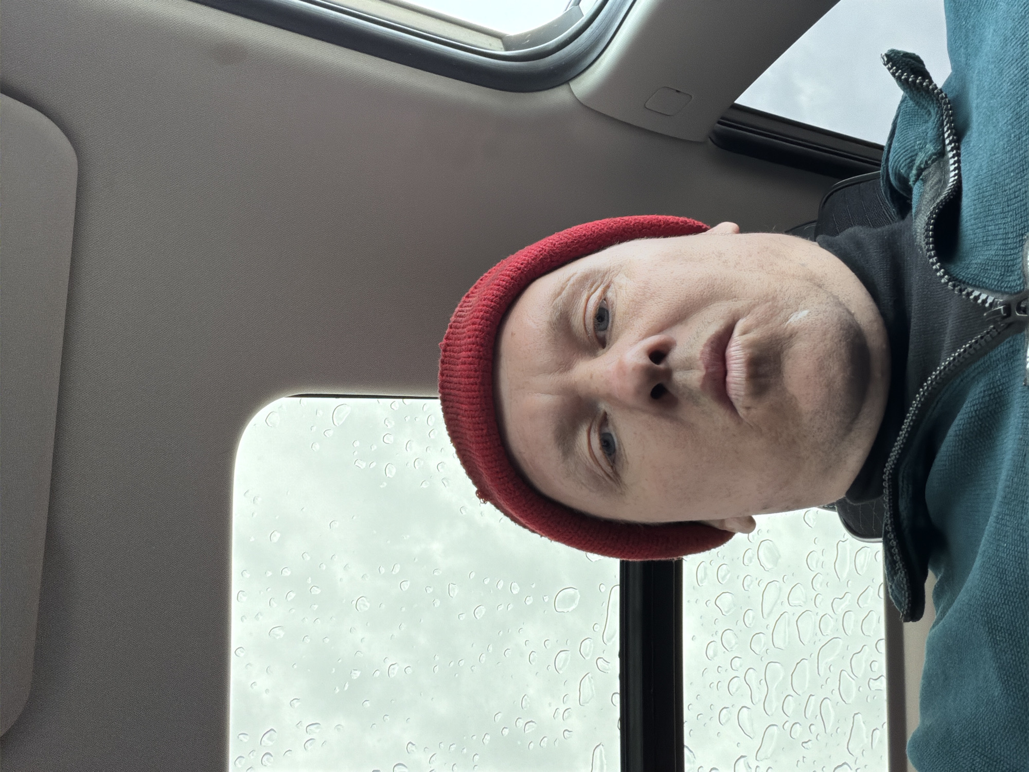 A selfie of a man wearing a red beanie and a blue jacket taken from inside a car during a rainy day. Water droplets are visible on the car window. The location is not discernible as Chaikovsky, Russia, based on the provided image.