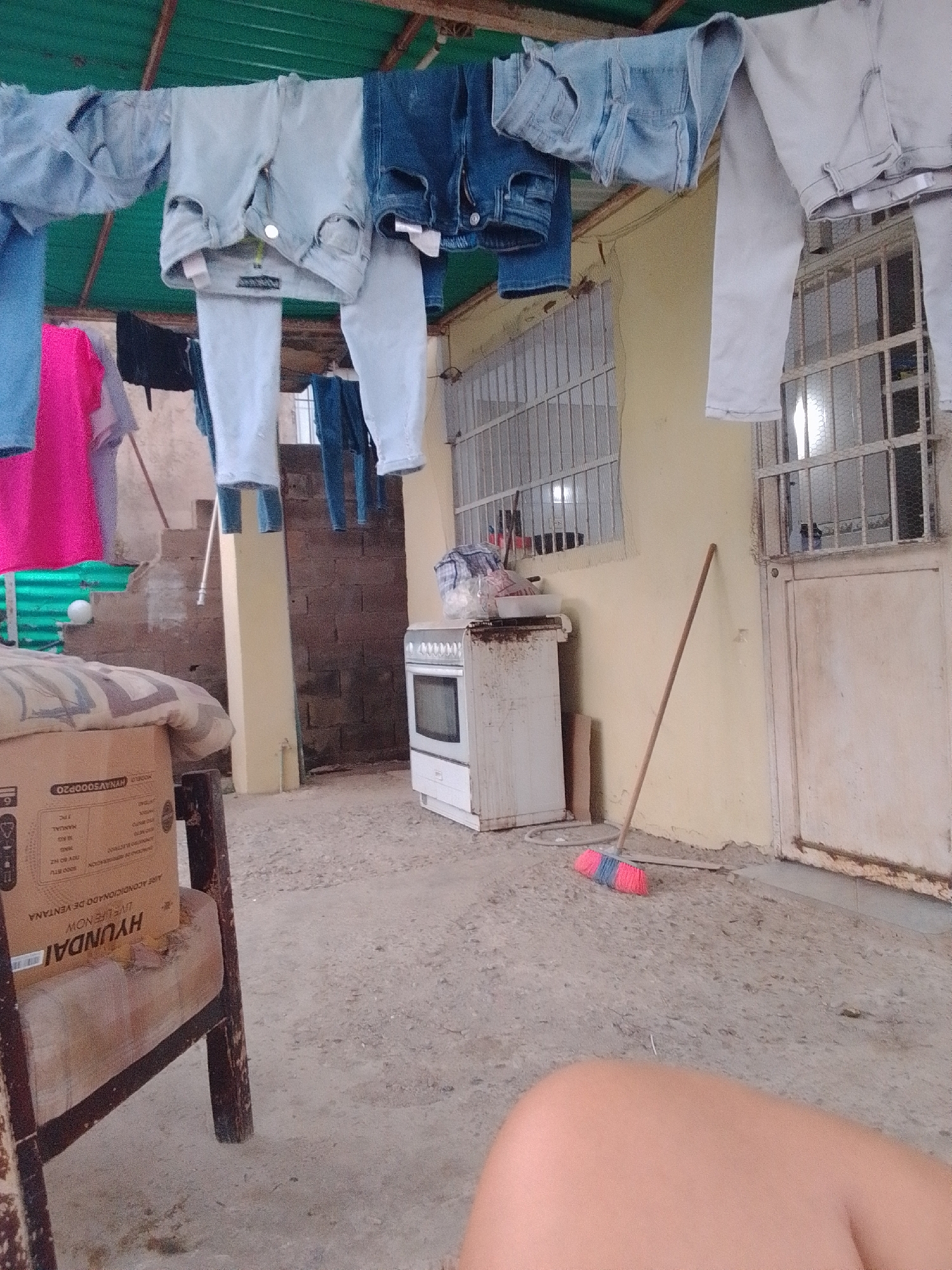 In La Tagua, Venezuela, an outdoor scene unfolds under a green corrugated metal roof, likely a patio or backyard. Laundry, primarily jeans and t-shirts in various shades of blue and pink, hangs on a clothesline, some pieces appearing damp. To the right, a narrow window with a metal grate looks into a building. In the background, a cream-colored oven with a visible oven door sits near a metal fence. A broom with a pink and blue brush leans against the wall next to the oven. A portion of a wooden chair and a cardboard box are visible on the left side of the frame, suggesting a somewhat cluttered or utilitarian space. The ground appears to be concrete or dirt.