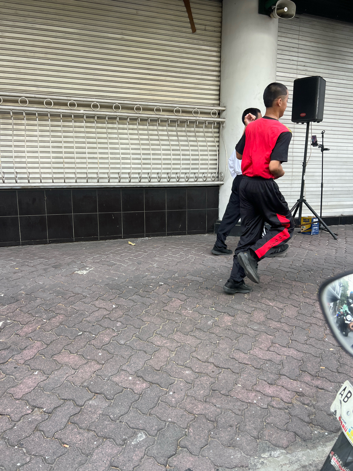 In a city setting, two young males are captured in motion on a brick-paved sidewalk. The individual in the foreground, wearing a red vest over a black t-shirt and black pants with red accents, is in mid-stride, his left leg extended forward. Beside him, another male, dressed in a white t-shirt and black pants, is also running. Behind them, a black speaker sits atop a tripod, with a microphone stand and a smartphone attached. A loudspeaker is mounted high on a white pillar to the left, and a closed metal roll-up door forms the background. The scene is illuminated by daylight, casting minimal shadows. The image appears to have been taken from a moving vehicle, as evidenced by the partial reflection of a motorcycle mirror in the lower right corner. The location is identified as Yogyakarta, Indonesia, suggested by the visible license plate.