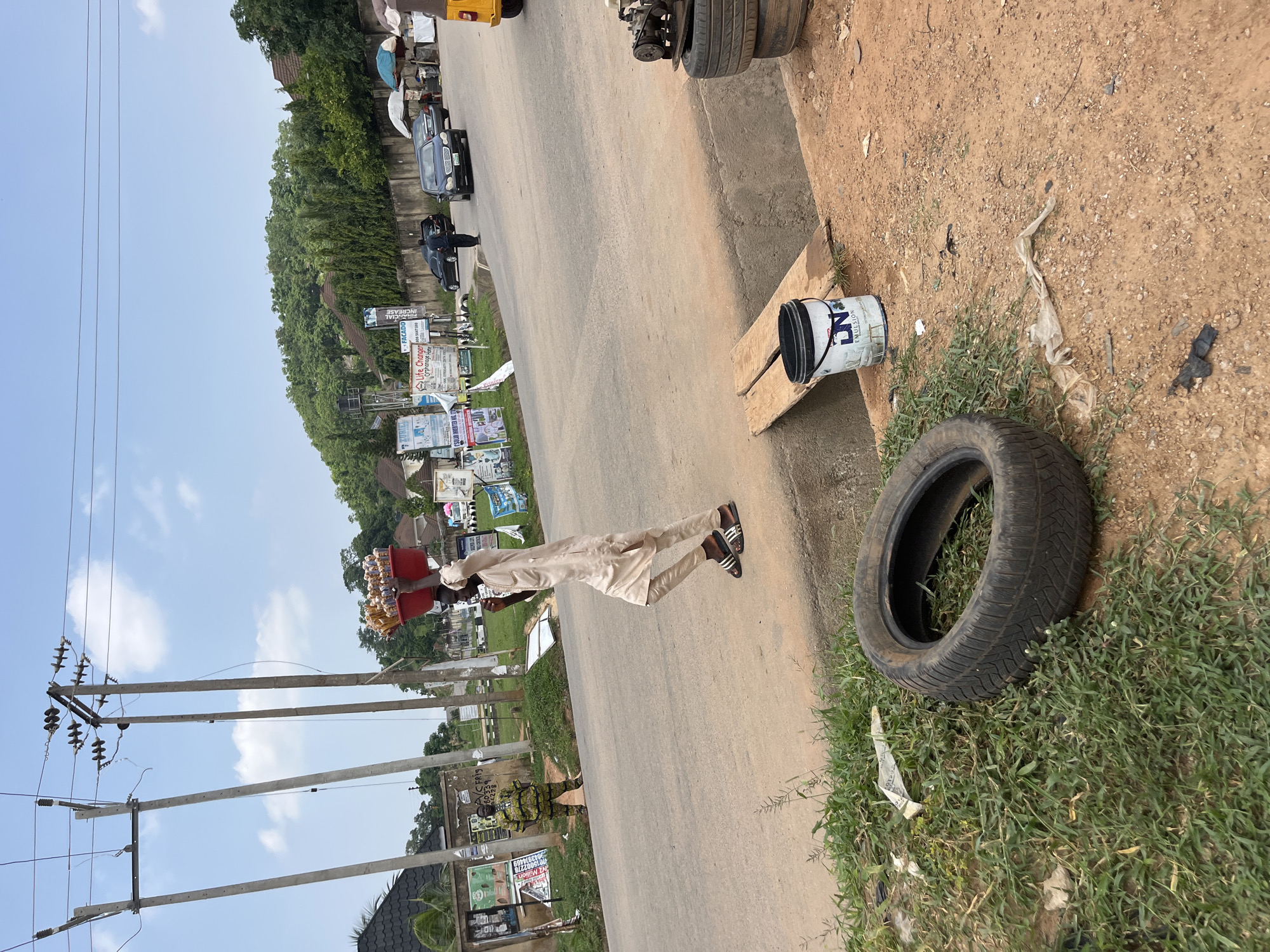 This is a vertical, eye-level, medium shot of a street scene in what appears to be Abuja, Nigeria. The road is paved, with a dirt and grass embankment on the right side. Several power poles with electrical wires are prominent in the foreground and extending into the background on the left. A person dressed in a light-colored traditional garment is walking on the road, carrying a red basket filled with yellow items, possibly food or snacks. They are wearing sandals.
On the right side of the road, a worn tire rests on the grass next to a wooden plank. A white bucket with a blue lid, labeled "LN EMULSION," is also on the plank. In the background, a line of trees forms a dense green backdrop. Beyond the trees, structures and more power poles are visible. There are various advertisements and signs placed along the side of the road. One sign reads "Life Changer Orphanage Home" and another displays "FACADO." The sky is mostly clear with some scattered clouds, suggesting it is daytime. The overall impression is a busy street with signs of commerce and everyday life. The presence of electrical infrastructure and the type of signage suggest an urban or semi-urban environment.