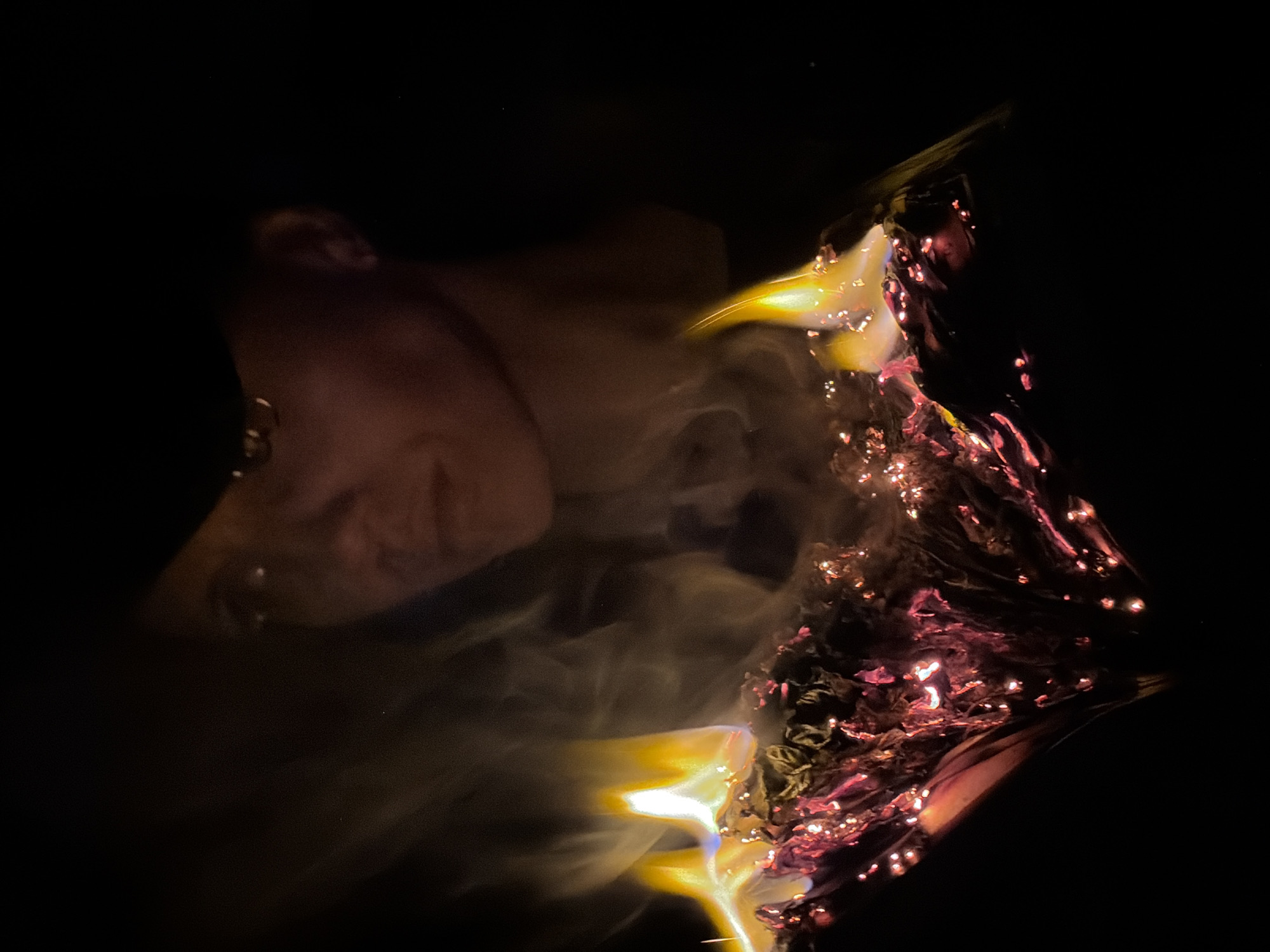 A close-up, low-light photograph shows a person's face partially visible on the left side of the frame, and an abstract fiery, smoky object dominating the right side. The person's face is blurred and lit with a warm, reddish-brown light. The object on the right appears to be in the process of burning, with bright yellow flames at its edges and embers glowing within a dark, swirling mass that appears to be smoke and the object itself. There are small red, bright sparks scattered throughout the object. The background is mostly black, with very little to no detail. The photo does not provide any information that would indicate the location is the city of Chaikovsky, Russia.