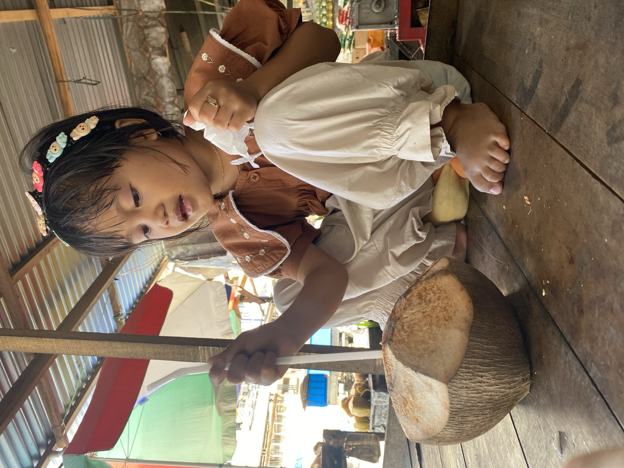 A young Southeast Asian girl with dark hair and a floral headband is sitting on a wooden surface, holding a straw and a piece of tissue. She is wearing a brown top with white trim and beige pants. In front of her is a halved coconut. The scene appears to be an outdoor market stall with a corrugated metal roof overhead and colorful umbrellas visible in the background. The overall impression is of a casual, everyday moment in a local Indonesian market.