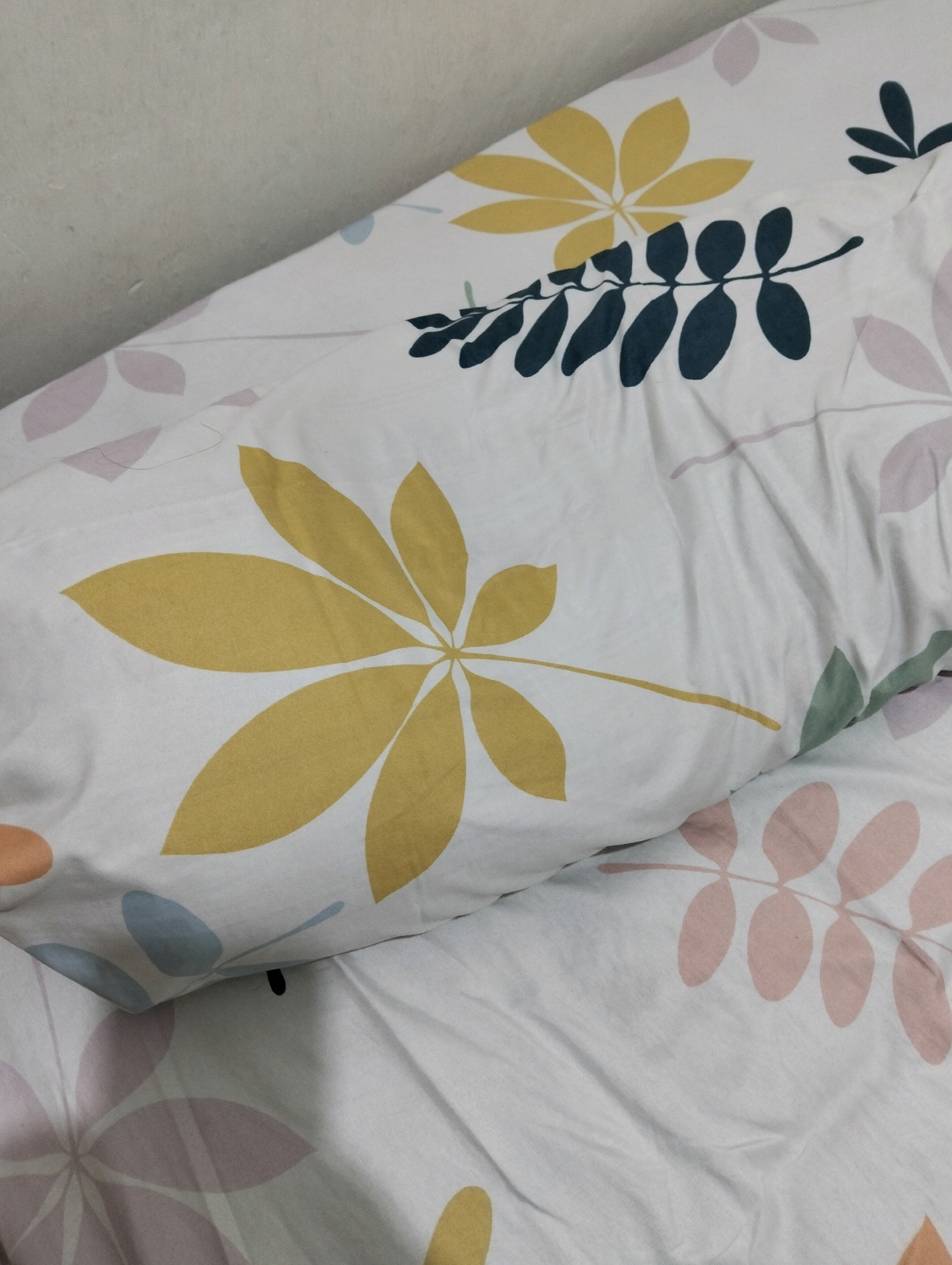 The image shows a close-up of a pillow and bedding with a floral pattern. The pillow is a light color with large yellow leaf-like shapes and smaller dark green leaf shapes arranged in a branch-like pattern. The bedding underneath also features a similar floral pattern with pink and light blue leaf shapes. The fabric is creased, indicating it is likely in use. The background is a plain, textured wall. The image appears to be taken indoors, likely in a bedroom. There is no information in the image to suggest a location in Malang, Indonesia.