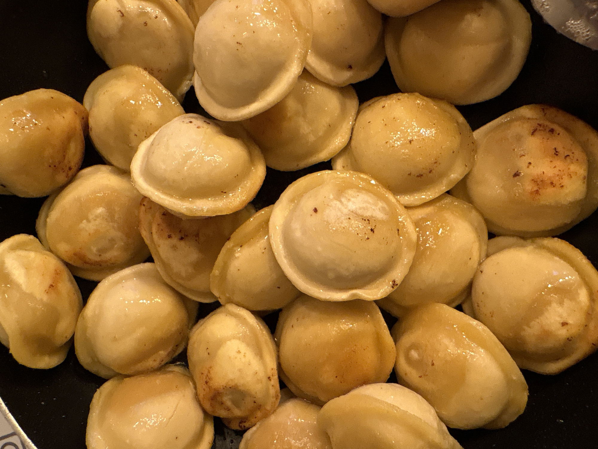 This media file is a close-up, top-down shot of numerous cooked pelmeni, a type of Russian dumpling, piled together in a black pan. The pelmeni are golden brown and appear slightly oily, with some showing a hint of darker browning on their edges, suggesting they have been pan-fried or boiled and then sautéed. Their surface is smooth and slightly glossy. The scene is a domestic setting, likely a kitchen, as indicated by the simple cooking vessel. There are no people visible, and the focus is solely on the food. The lighting appears to be artificial, common for indoor food photography. The context of Krasnoyarsk, Russia, places this dish within its cultural origin. There is no visible text in the image. The overall impression is one of a hearty, prepared meal.