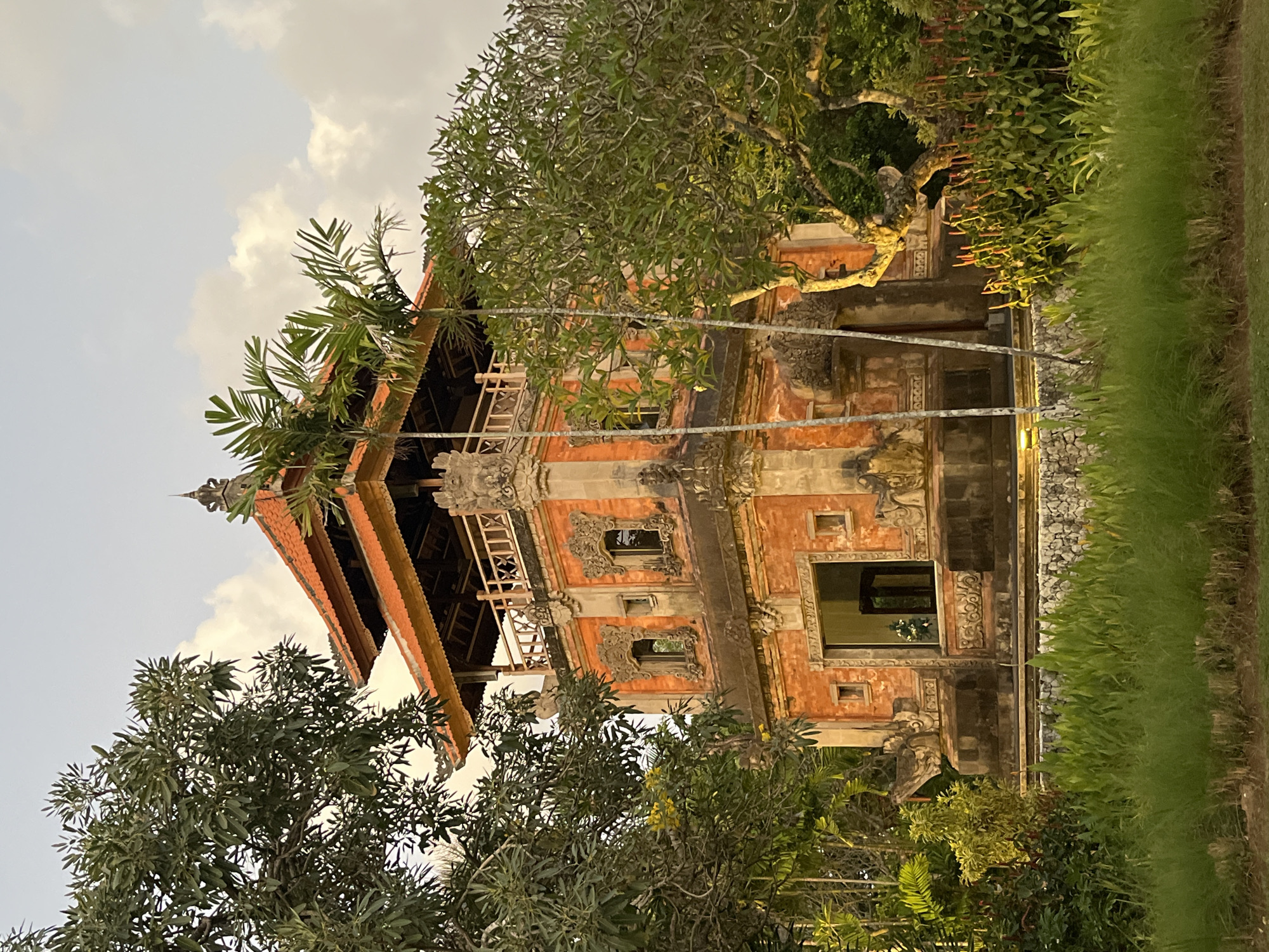 The image depicts a multi-story Balinese-style building with orange brick walls and ornate carvings, set amidst lush greenery and tall trees. The building features a tiered, terracotta-tiled roof, wooden balconies, and decorative window frames. The foreground is filled with vibrant green grass and foliage. In the background, the sky is overcast with soft clouds. The architecture suggests a tropical location, likely a temple or a traditional residence in Bali, Indonesia. The lighting indicates it is either early morning or late afternoon. No people are visible in the image.