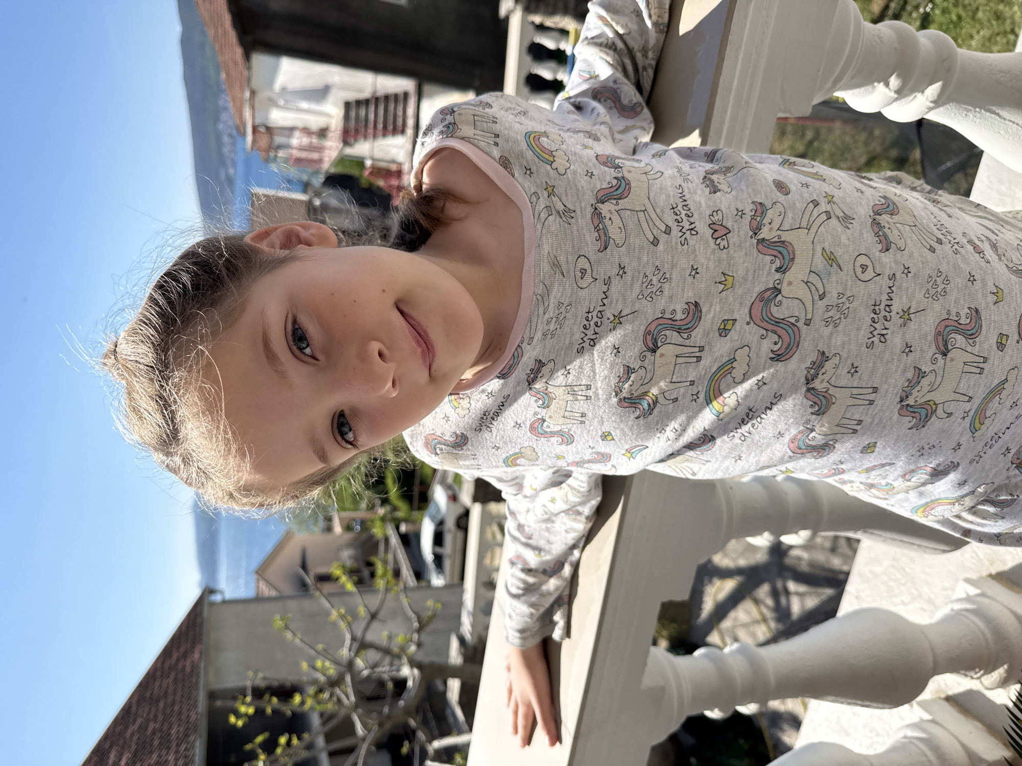 A young girl with blonde hair and blue eyes is leaning on a white balcony railing. She is wearing a light gray, long-sleeved shirt with a unicorn and rainbow pattern. The background shows a cityscape with white buildings, a blue sky, and a body of water. The location is Bijela, Montenegro.