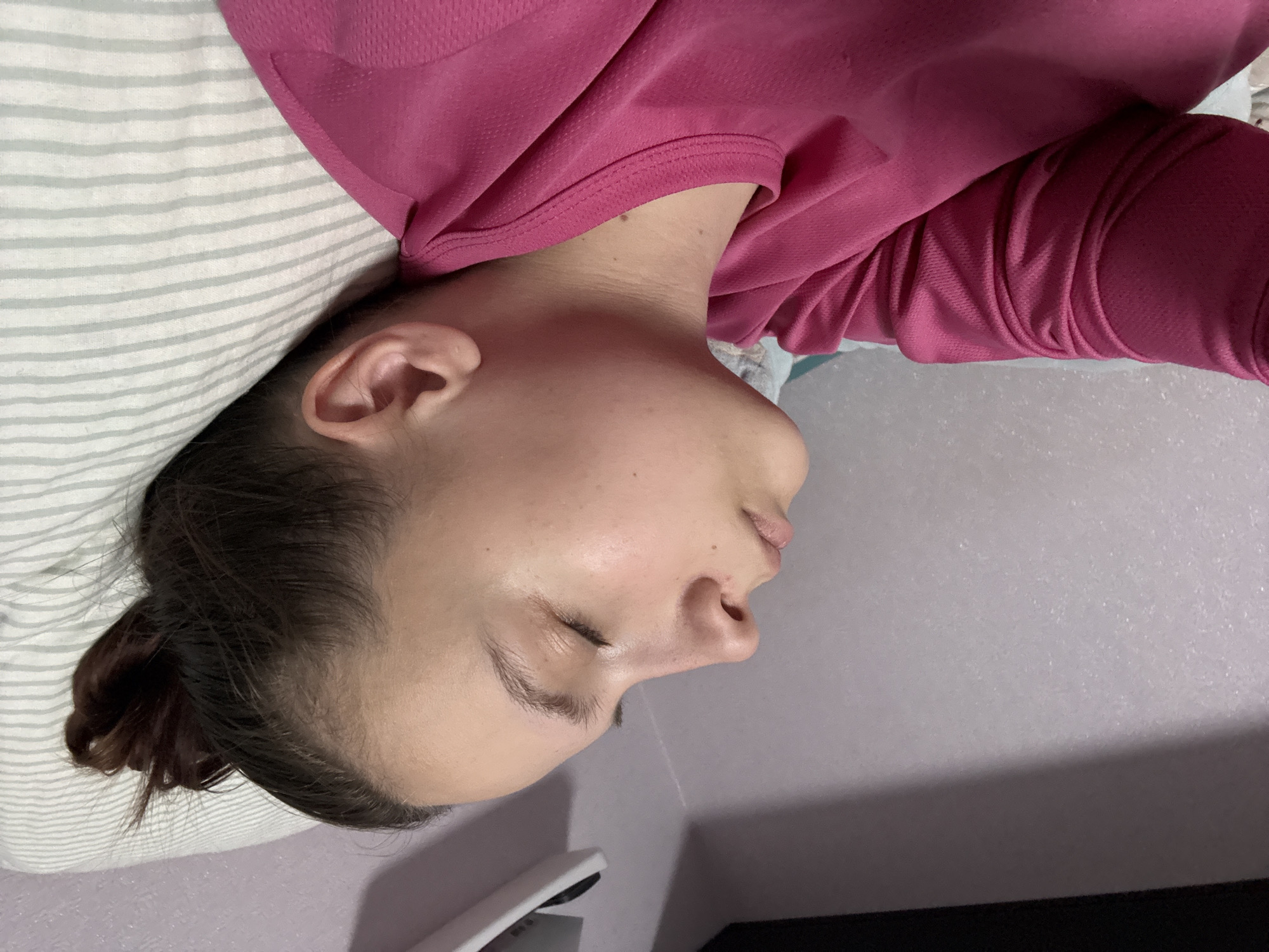 A person lies on their side, possibly sleeping, on a striped pillow. They are wearing a pink long-sleeved shirt. Their hair is pulled back. The location is unknown, and there is no verifiable connection to Прикамский, Russia.