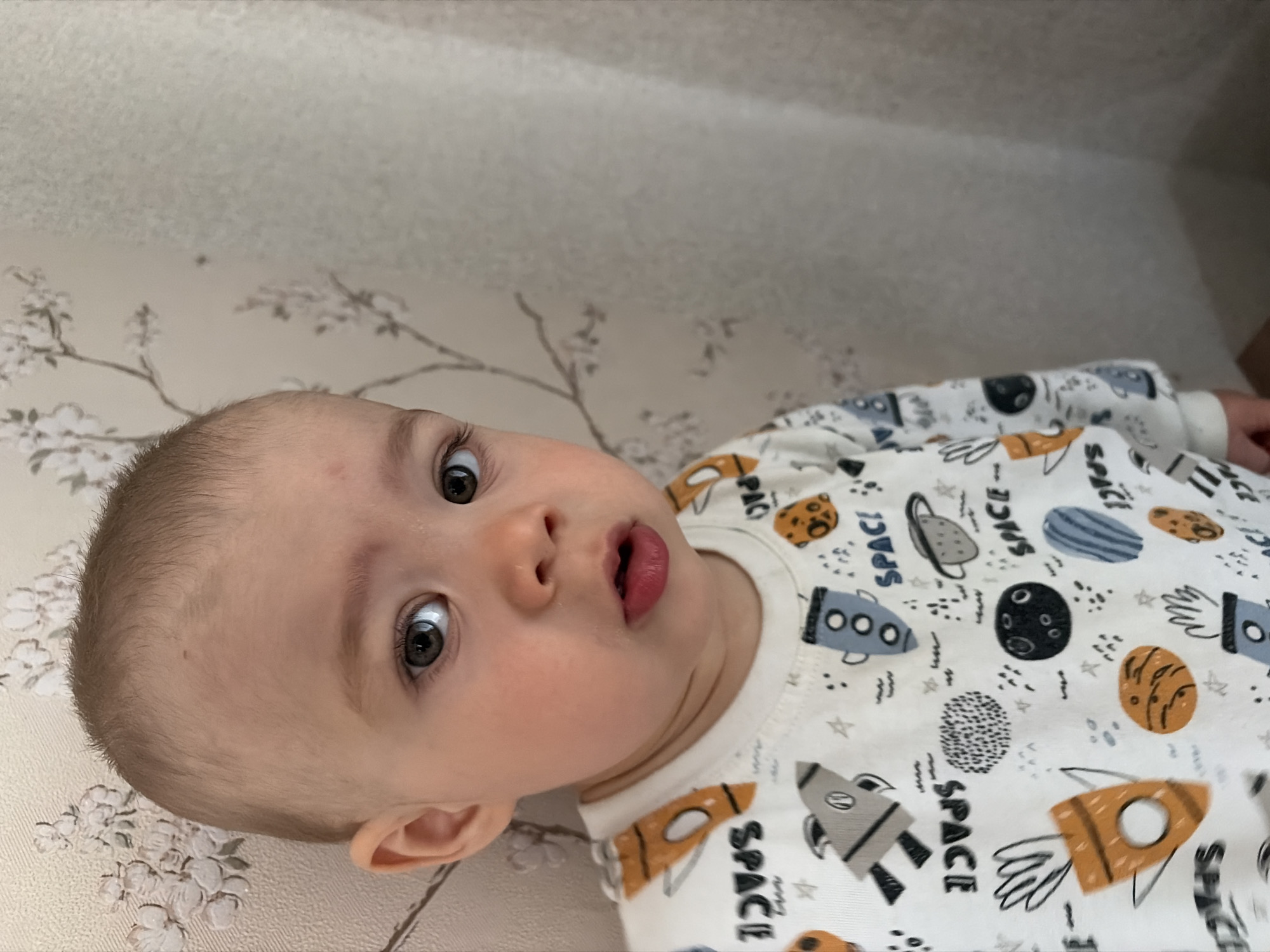 An infant is captured in a close-up, lying on their back and looking directly at the camera with wide, light-colored eyes and a slightly open mouth, conveying a sense of curiosity or mild surprise. The baby has fine, light hair and distinct facial features.
The infant is dressed in a white long-sleeved top featuring a playful space-themed pattern. The design includes numerous rockets in orange, grey, and blue, alongside stylized planets and celestial bodies depicted in black, orange, and blue striped, and speckled motifs. Small stars are scattered across the fabric, and the word "SPACE" is visible multiple times, printed in a simple font, adding to the theme.
The background consists of a light-toned surface, possibly wallpaper or bedding, adorned with a delicate, lighter-colored floral or branching pattern. This suggests an indoor setting. The photo is taken from a slightly overhead perspective, with the baby's head positioned towards the left side of the frame. While the location context is Chaikovsky, Russia, no specific visual cues within the image itself indicate this geographical detail. The lighting appears soft and even, highlighting the baby's features and the details of their clothing.