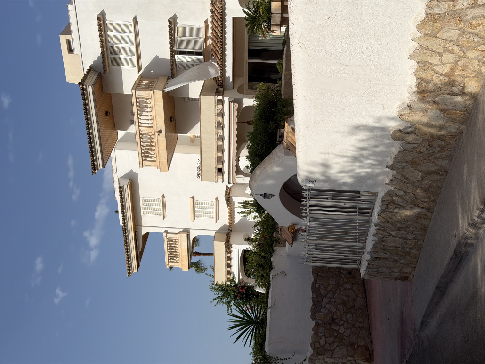 The image shows a white multi-story building with beige balconies and terracotta roof tiles in Calvià, Spain. The building has several windows with white shutters. Lush green plants and a palm tree are visible around the base of the building and on the balconies. A white wall, partly constructed with rough stones, encloses the property in the foreground. A metal gate with the number "18" is visible on the wall. The sky is blue with some scattered clouds. The scene appears to be a residential building in a sunny, Mediterranean climate.