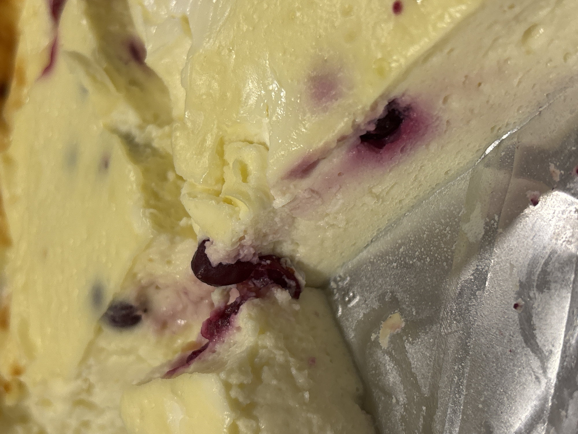 A close-up shot depicts a section of what appears to be a blueberry cheesecake. The dessert is light yellow in color with visible purple blueberry inclusions. The texture of the cake is soft and creamy. Part of the dessert is enclosed in a silvery, rough-textured container, likely a foil wrapper. The lighting is diffused, creating soft highlights on the creamy surface of the cake. There's no information to suggest that this image was taken in Perm, Russia. There are no people or subjects beyond the food item visible in the image. The main action is the presentation of the dessert, though no interaction or environment beyond the container is apparent.