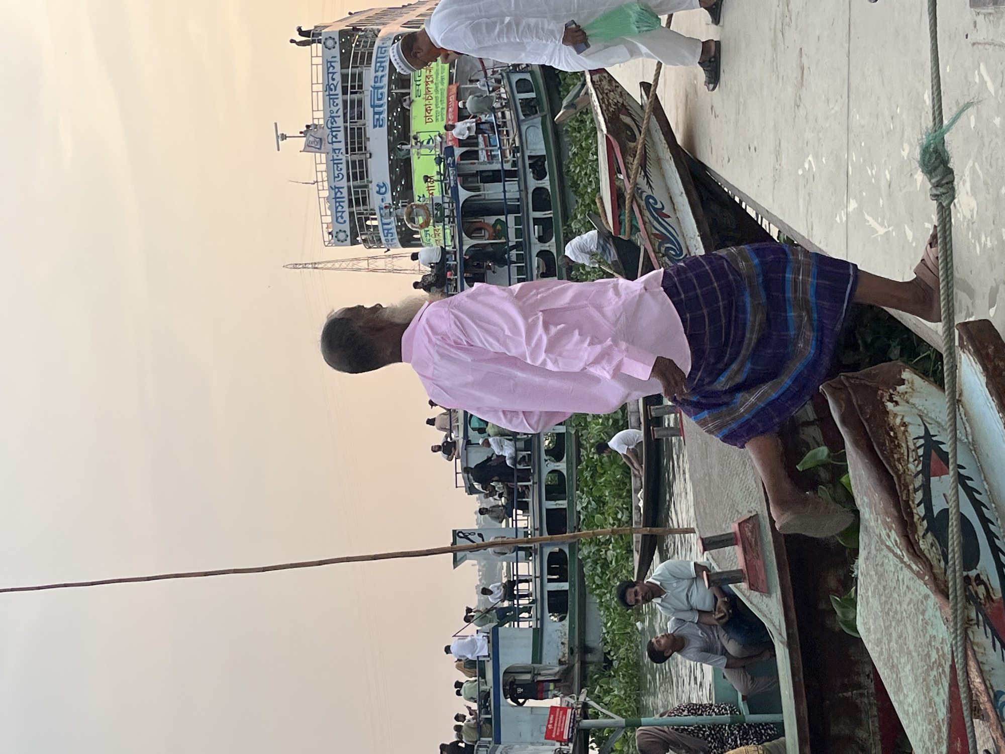 This photo, taken in Dhaka, Bangladesh, depicts a man in a pink shirt and purple lungi standing in a narrow boat, possibly a sampan, near a larger ferry. The ferry, named "Messrs. Dollar Shipping Lines," appears crowded with passengers. The environment suggests a river or waterway setting in Dhaka.