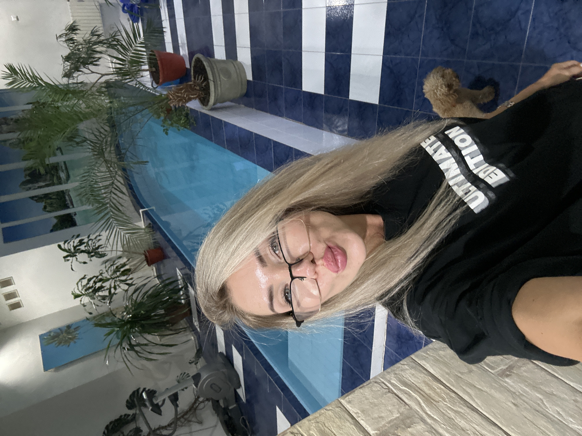 A woman with blonde hair, wearing glasses and a black t-shirt with "ULTIMATE EDITION" printed on it, takes a selfie in front of a swimming pool. She has her lips puckered. A small, fluffy, light brown dog stands to her right. The environment appears to be an indoor pool area with tiled flooring in a blue and white striped pattern, and several potted plants around the pool. The woman is positioned in the foreground, with the pool and plants in the background. The image is a snapshot of a personal moment. There is no information in the image to confirm its location in Armavir, Russia.