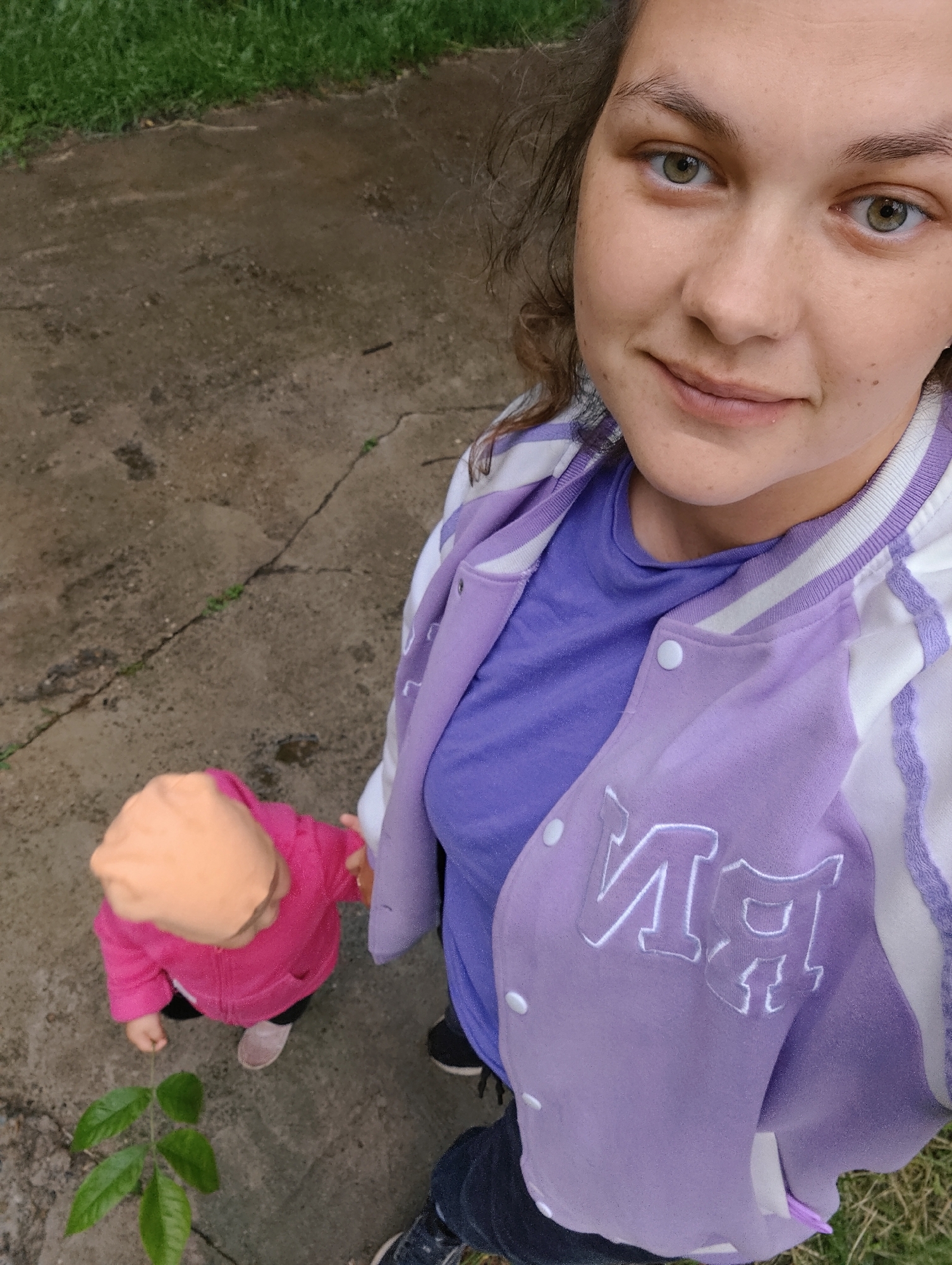 A high-angle outdoor selfie captures a woman and a small child in Chaikovsky, Russia. The woman, prominently in the upper right, has curly brown hair, green eyes, and a light complexion with freckles, offering a gentle smile. She is wearing a purple t-shirt beneath a lavender and white varsity-style jacket with white buttons and striped cuffs. The Cyrillic letters "ИЯ" are clearly embroidered in white on her jacket.
Below her and to the left, a small child is seen from an overhead perspective, wearing an orange or peach-colored hat/hood, a bright pink zip-up jacket, and light-colored shoes. The child is looking down intently at a small green plant with distinct leaves growing on the ground. The setting is a cracked, damp concrete surface with patches of green grass visible along its edges. The scene takes place outdoors during the daytime, with the wet ground suggesting recent rain or an overcast sky. The image conveys a tender, everyday moment between the woman and child.