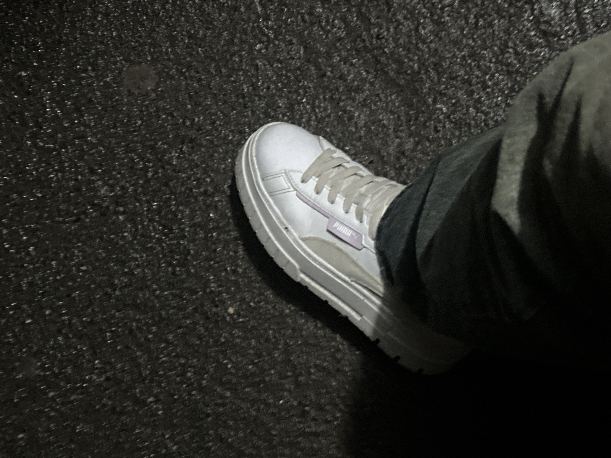 This is a low-angle, full-shot, eye-level photograph of a person's left foot wearing a white Puma sneaker with grey laces and purple accents. The shoe is placed on a dark, textured asphalt surface that appears wet, suggesting it might be raining or has recently rained. The background is predominantly the dark asphalt, with some lighter speckles that could be reflections or debris. The lighting is dim, casting a shadow to the left of the shoe and obscuring the lower portion of the person's leg, which is covered by dark-colored pants. No specific landmarks in Togliatti, Russia are visible. The scene is devoid of any other people or discernible activity.