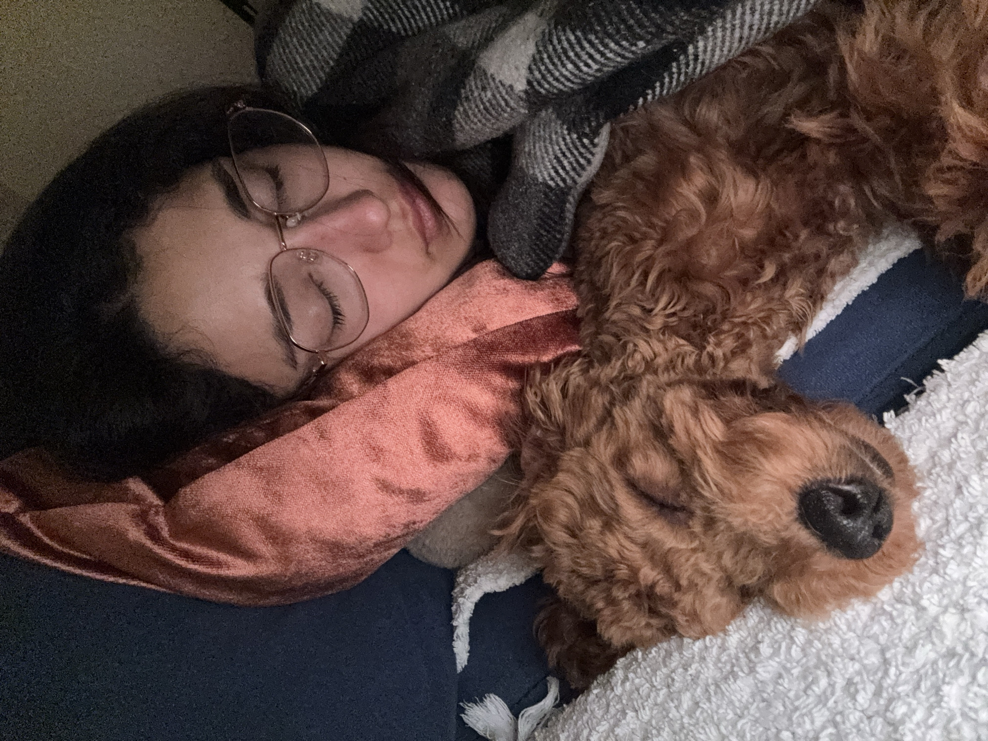 The image shows a woman and a dog sleeping together on a couch. The woman is on her left side, wearing glasses, with a peach-colored scarf around her neck and a gray and black blanket partially covering her head. The dog, a curly-haired breed (likely a doodle or similar), is nestled against her on top of a white towel. The couch has dark blue upholstery. The location in the city of Sutomore, Montenegro is not evident from the visual information provided.