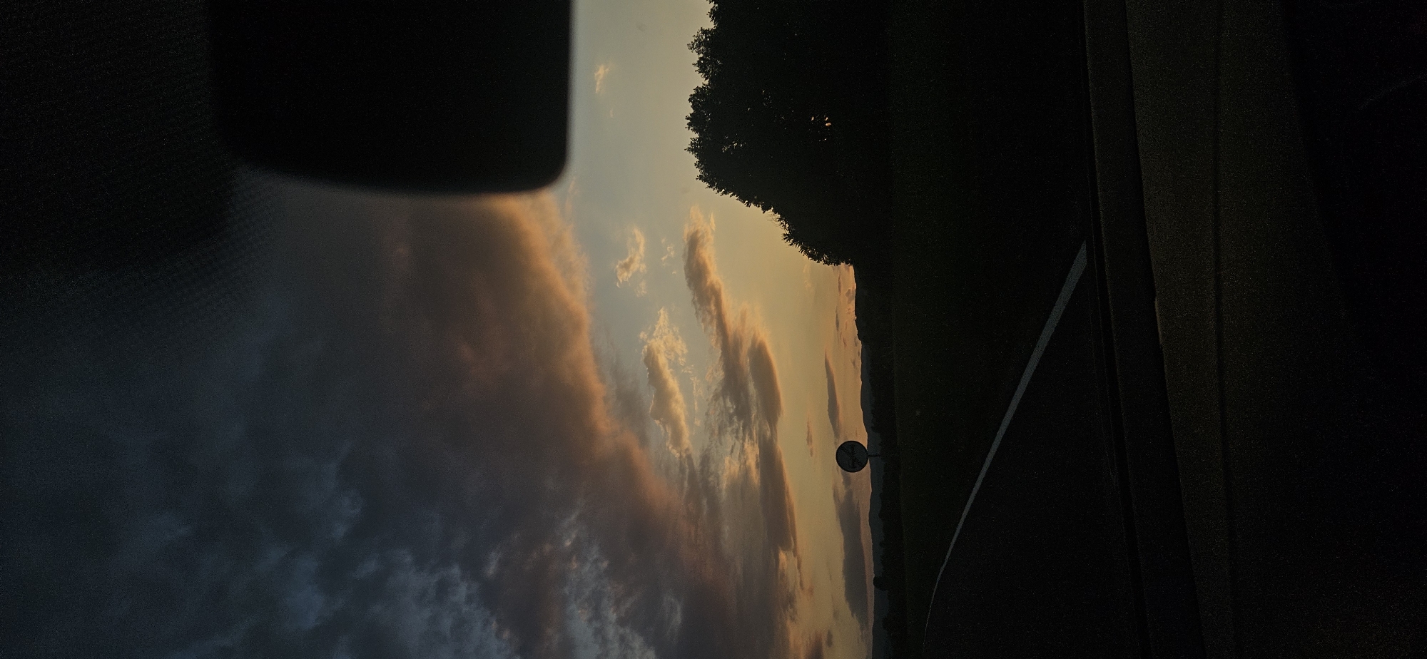 The image captures a sunset landscape as viewed from inside a moving vehicle, located in the city of Dolna banya, Bulgaria. The upper portion of the frame is dominated by a twilight sky, transitioning from dark blue-grey, heavy clouds in the upper left to vibrant orange, yellow, and peach hues among lighter clouds towards the horizon on the right, indicating a setting sun.
In the immediate foreground, the dark silhouette of a vehicle's dashboard or interior is visible along the bottom edge. A white solid line marks the edge of a paved road, which curves gently towards the left in the lower-left quadrant. To the right of the road, the landscape consists of dark, silhouetted fields and a dense line of trees forming a dark mass against the illuminated sky in the mid-right. A distinct, round road sign with a pole is visible in the mid-ground, centered horizontally, and appears largely silhouetted, obscuring its specific symbol. Distant hills or mountains form a faint, dark silhouette against the horizon beneath the sunset clouds. A dark, rectangular object, possibly a rearview mirror or part of the car's interior roof lining, intrudes into the upper-right corner of the frame. No individuals are present in the scene, and no specific actions or interactions are depicted.