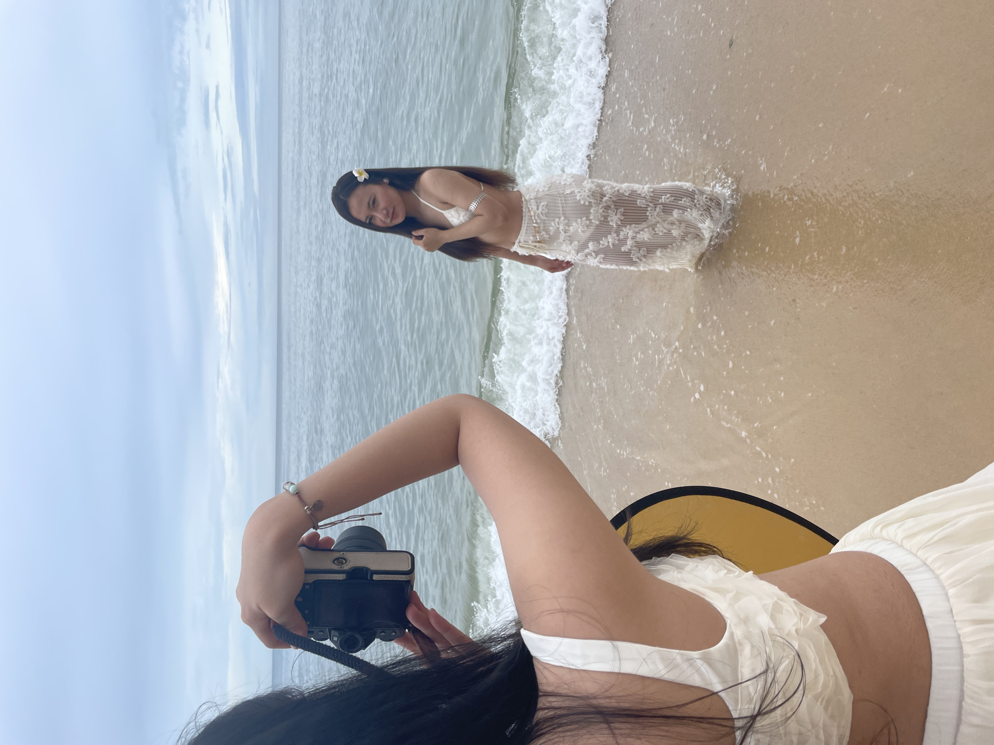A low-angle, outdoor shot captures a woman in a white bikini top and white lace skirt standing in the ocean surf on a beach. The sky is overcast and the ocean is gray. Another woman is partially visible in the foreground, holding a black and gray camera and wearing a white frilly top and white skirt. A yellow reflector is positioned near the photographer. The location is likely Phu Thuy Ward, Vietnam, based on the likely location.