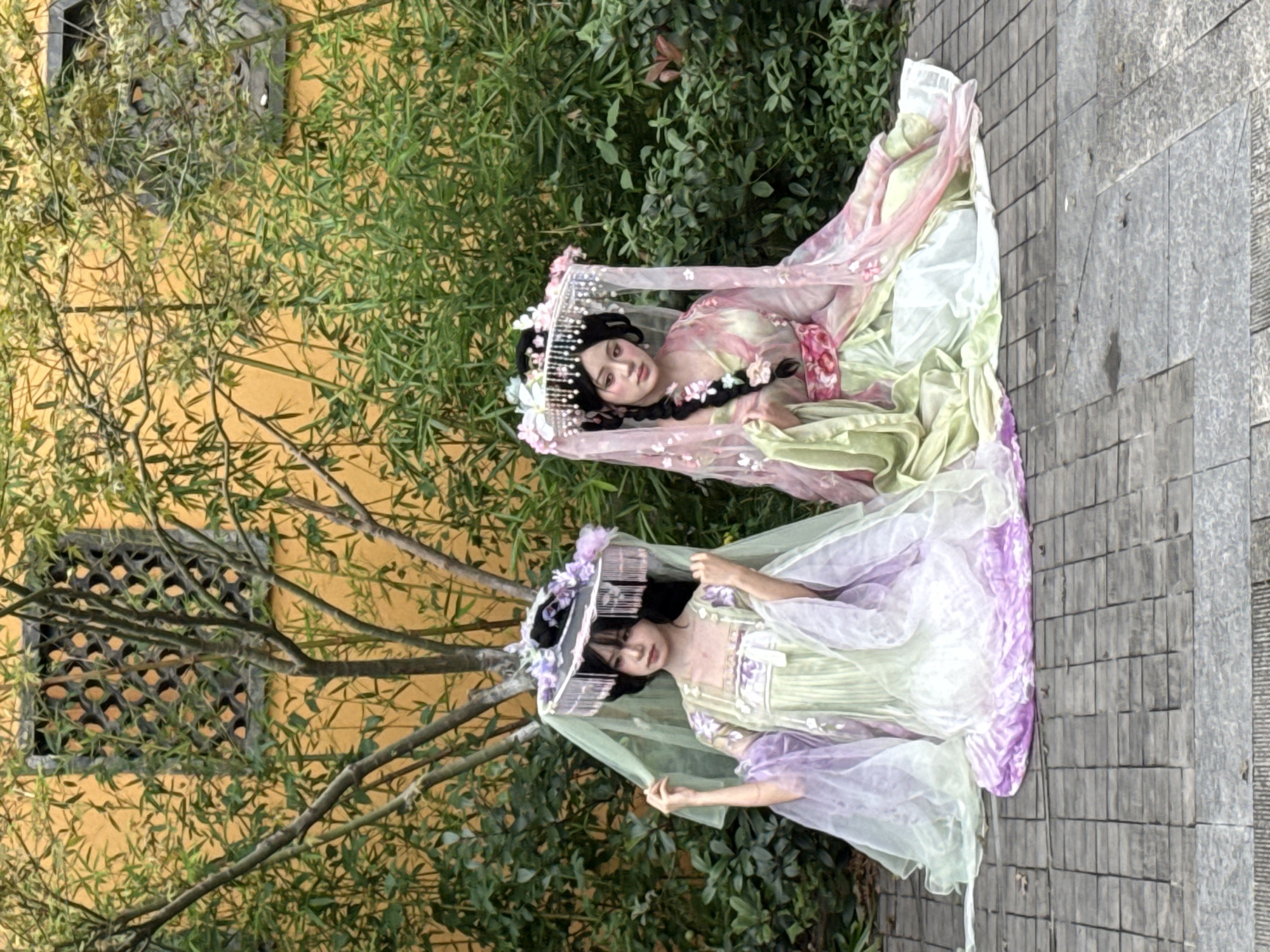 Two young women in traditional Chinese Hanfu dresses are sitting on a stone pathway in front of a yellow wall and a bamboo grove. The woman on the left wears a light green and lavender dress with a decorative headdress. The woman on the right wears a light green and pink dress, also with an ornate headdress. Both women have dark hair and are looking towards the camera. The setting appears to be a garden or park. The specific location in Yuzhong District, China, cannot be determined from the image alone.