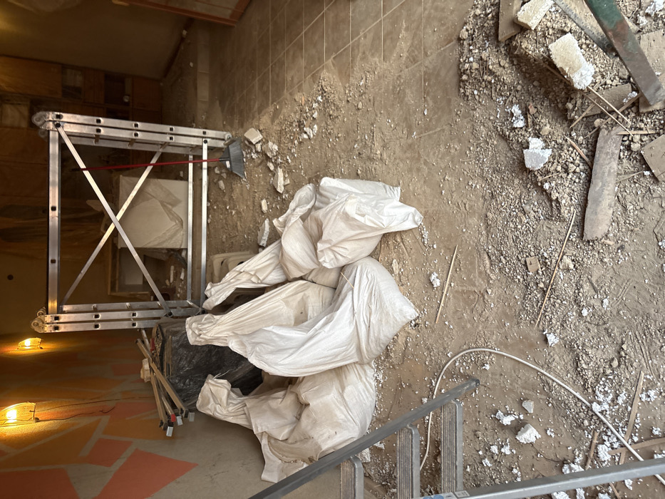 The image displays an indoor space in Stefan Karadja, Bulgaria, undergoing renovation or demolition. The floor is primarily composed of loose dirt, gravel, and construction debris, including fragments of concrete, plaster, and wood splinters. Approximately seven large, white, opaque bags, appearing to contain debris or construction waste, are piled in the central foreground. To the immediate left, the lower section of an aluminum step ladder is visible, with a white electrical cable coiled on the ground nearby. The left wall features an abstract, mosaic-like pattern in shades of orange and brown, illuminated by two exposed incandescent light bulbs.
In the midground, a substantial, silver-colored aluminum collapsible scaffolding unit is erected, with diagonal cross-braces providing structural support. A red-handled broom leans against the right side of this scaffolding. Behind the scaffolding, a large, light-colored object, possibly wrapped or covered, is present. The floor on the right transitions into a section of light brown square tiles. The far right background reveals a partial view of a wooden door frame or panel. No individuals are present in the scene. The overall environment is dusty and disarranged, consistent with a construction site.