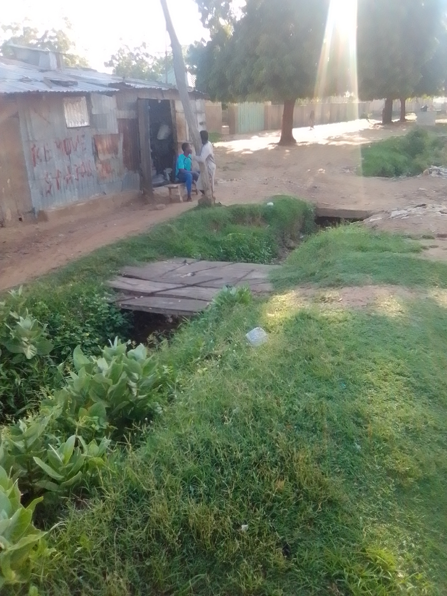In Kwadon, Nigeria, a scene unfolds near a corrugated iron structure. Two individuals are present; one, a young male wearing a blue t-shirt and dark trousers, sits on a wooden bench outside the structure. Standing beside him, dressed in a white garment, is an adult male. The structure appears to be a shop or dwelling, with red lettering visible on its exterior. A wooden footbridge spans a small, overgrown channel of water in the foreground, with lush green vegetation bordering the channel and extending into the foreground. The ground is a mix of dirt and sparse grass, with trees visible in the background, some illuminated by strong sunlight.