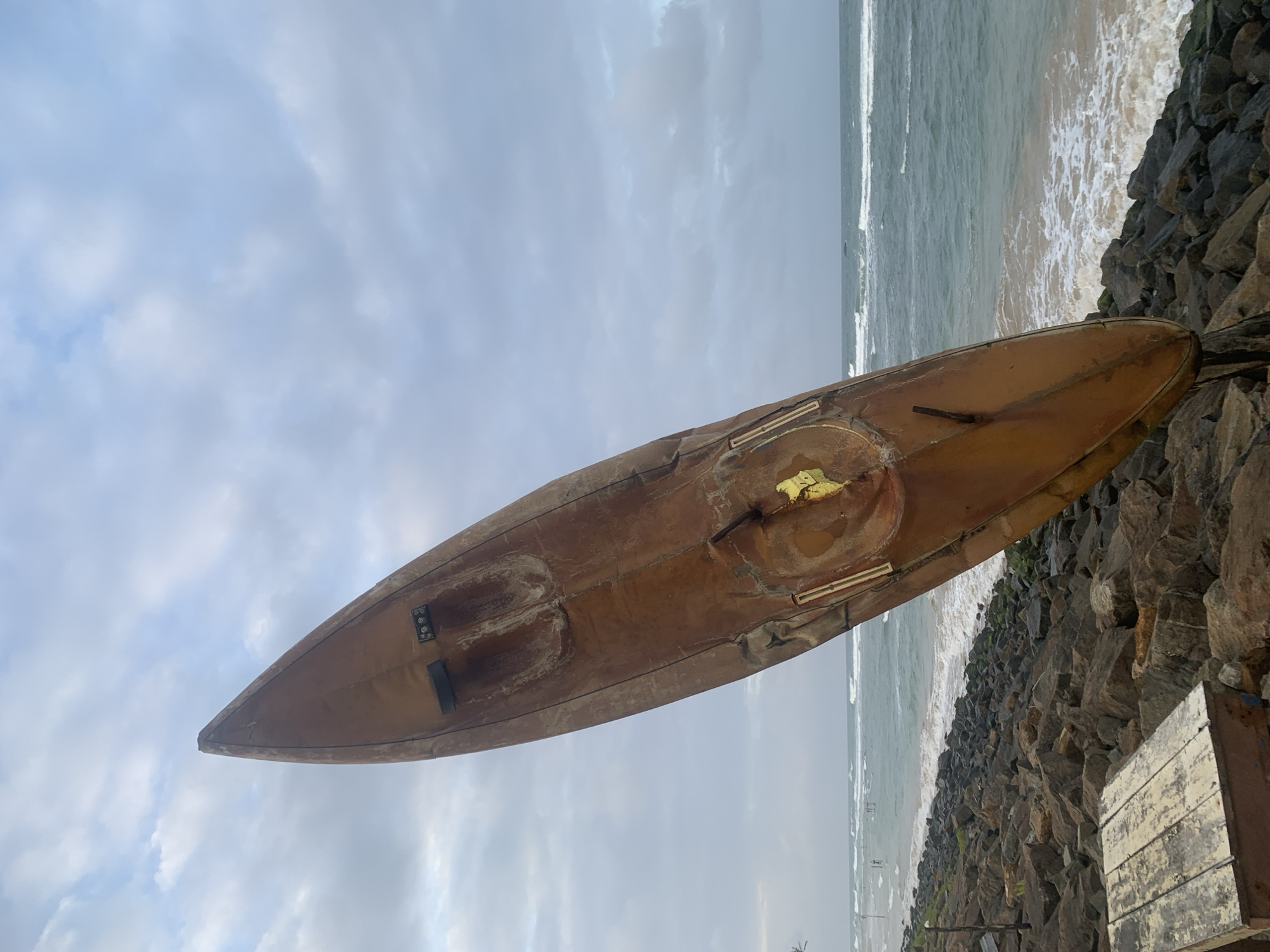 A weathered, rusty orange surfboard is propped upright on a rocky shoreline in Kathaluwa, Sri Lanka. The surfboard's pointed end faces the cloudy sky, while its base rests amongst large, dark stones. The ocean stretches out behind it, with waves crashing onto the shore. A wooden crate sits in the foreground, also on the rocks. The scene appears to be captured on an overcast day.