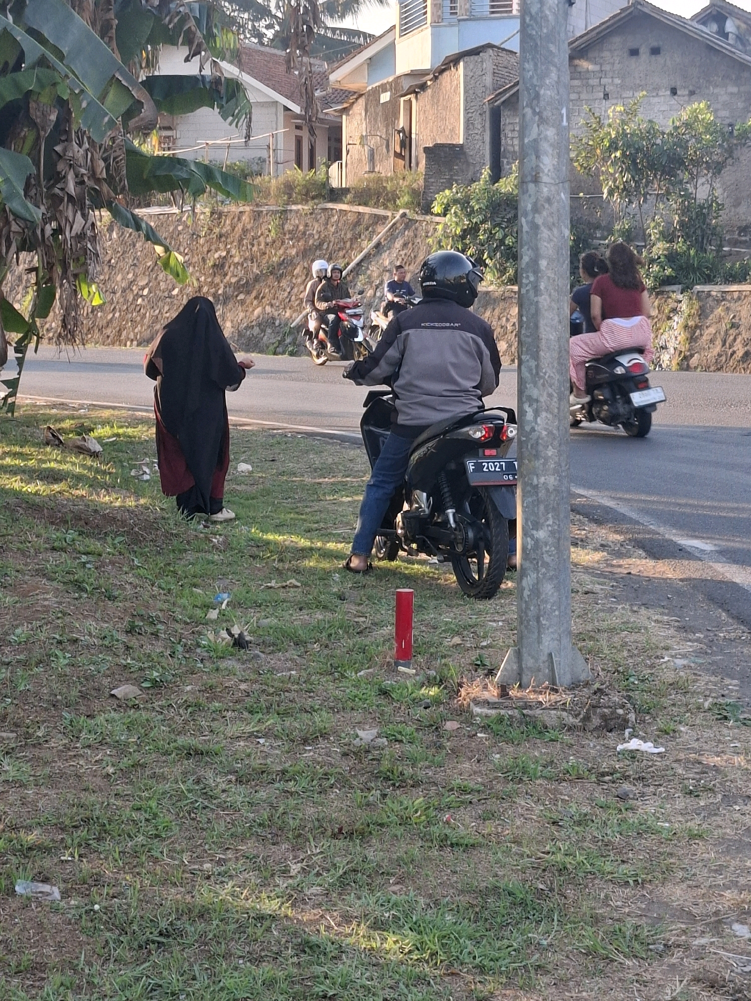 In Cikaret, Indonesia, on a paved road bordering a grassy embankment, a scene unfolds under natural daylight. A person in a black hijab and a maroon dress stands on the grass, facing away from the camera, near a banana tree. To the right, a man wearing a black helmet and a grey jacket rides a black motorcycle. Behind him, on the same motorcycle, a woman with a maroon shirt and striped pants is visible. Further down the road, another motorcycle carries two individuals, also facing away from the camera. Buildings with stone walls and tiled roofs line the background, with lush green foliage interspersed. A tall, grey utility pole stands prominently in the foreground. The overall environment suggests a rural or semi-urban setting.