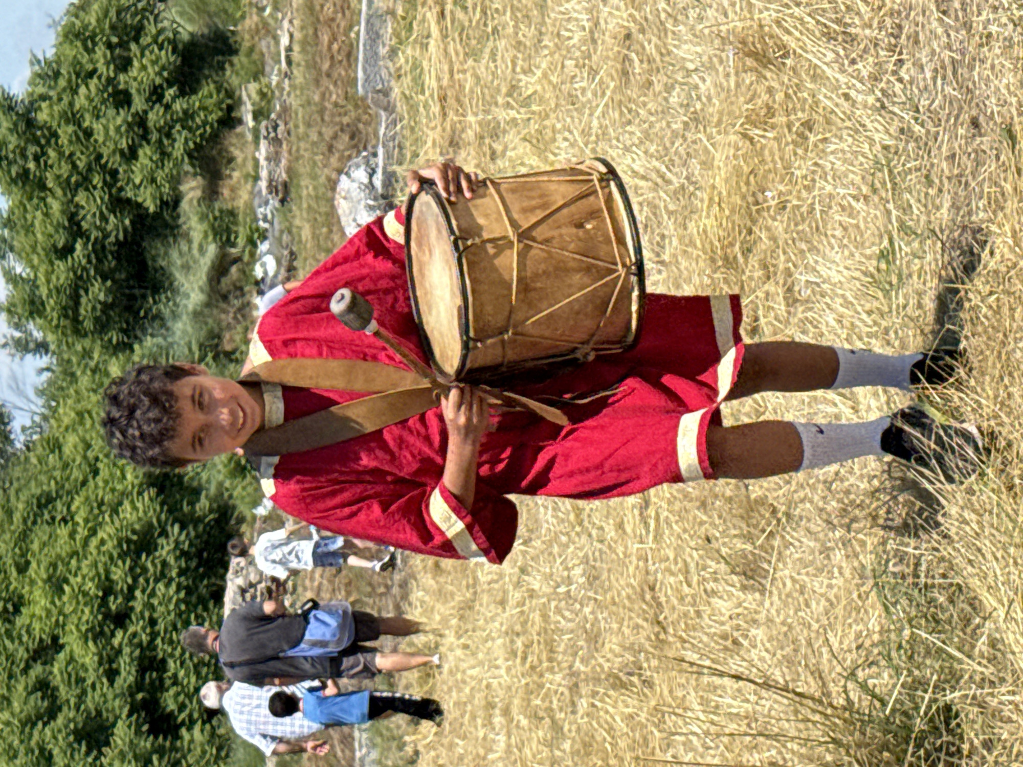 A young male, likely a pre-teen, is positioned in the foreground, facing the camera with a slight smile. He wears a red, knee-length tunic with light-colored trim at the cuffs and hem, and a brown strap diagonally across his chest. He has white socks and dark athletic shoes. He holds a cylindrical, light brown drum with a dark rim in his left hand, and a short drumstick in his right hand, poised above the drum's surface. The setting is an outdoor, sunlit area within the city of Gigen, Bulgaria. The ground is covered with dry, light-colored grass and straw. In the mid-ground, several individuals are visible: an adult male in a light checkered shirt, an adult male in a dark t-shirt carrying a blue bag, and a young boy in a blue t-shirt, walking from left to right. Another young boy in a white t-shirt and blue shorts is further to the right. The background features scattered stone remnants, indicative of an archaeological site, and dense green foliage under a bright sky.