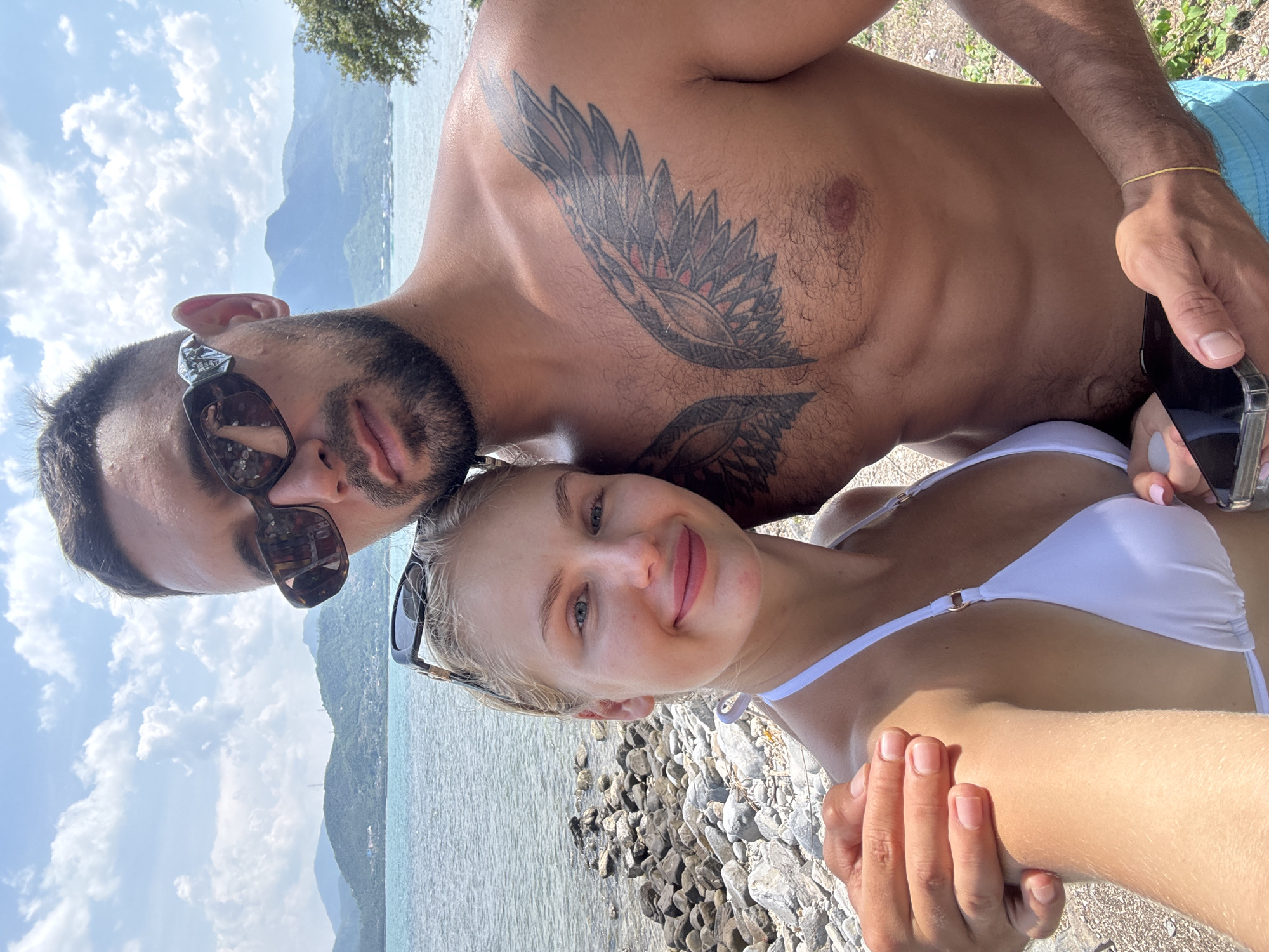 The image shows a selfie taken by a man and a woman on a beach. The man is shirtless, revealing a large tattoo of wings on his chest. He is wearing sunglasses and blue swim trunks. The woman is wearing a white bikini and sunglasses. They are standing close together, with the man's arm around the woman's shoulder. In the background, there is a body of water, a rocky shore, and mountains. The sky is blue with some clouds. The location is identified as Ninh Ích, Vietnam.