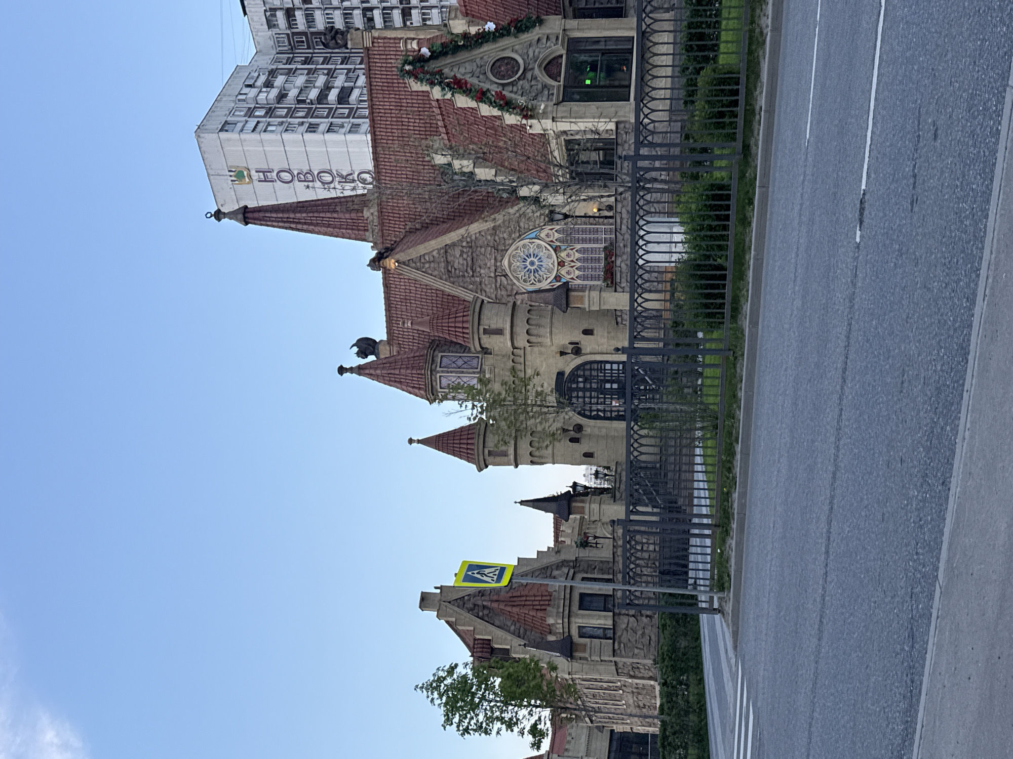 The image shows a street scene in Moscow, Russia, featuring a building designed in a fairytale castle style. The building has multiple turrets, stone walls, and a red-tiled roof. Above the castle-style building is a modern, multi-story building with a sign reading "HOBOKO" in Cyrillic. A black metal fence separates the building from the sidewalk and road. A pedestrian crossing sign is visible. The road is asphalt with white lane markings. The sky is clear and blue.