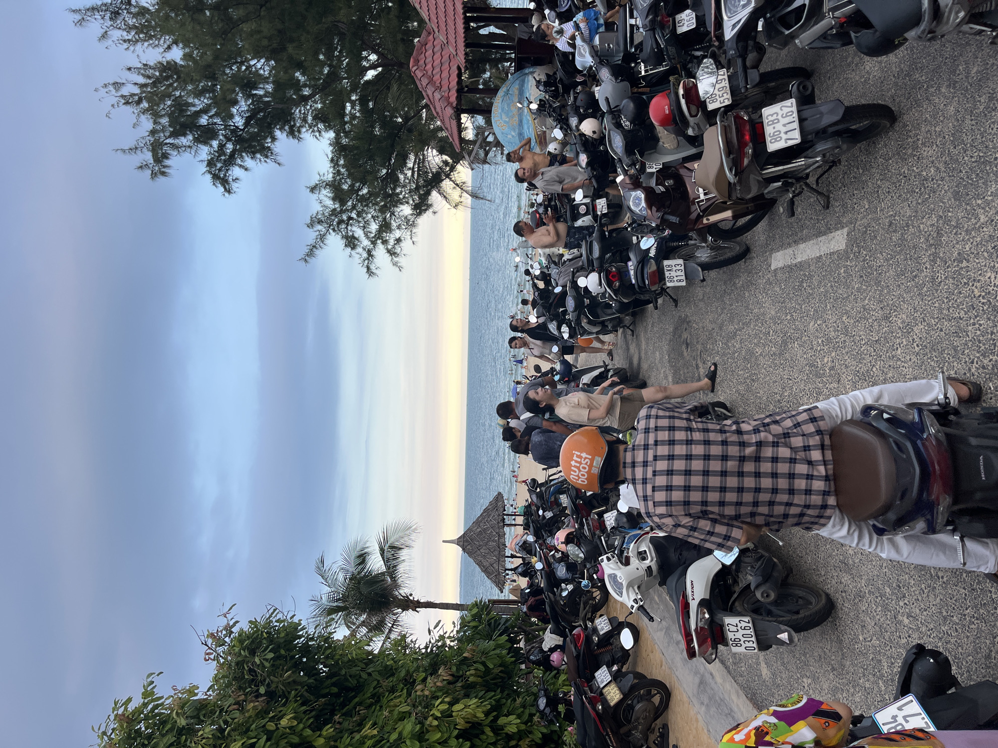 The image depicts a parking lot filled with numerous motorbikes situated adjacent to a beach. In the background, the ocean stretches towards the horizon under a cloudy sky. A few people are visible on the beach and near the water. The parking lot is paved and lined with motorcycles. Some people are standing and conversing among the parked vehicles, while others are seated on motorbikes. A man in a checkered shirt and white pants is seated on a motorbike in the foreground, facing away from the camera. Another person on a motorbike to the left of the frame is partially visible. Trees and foliage line the left side of the scene, and a thatched-roof structure with red roofing stands near the beach. The scene suggests a popular public area, likely a beach in Phan Thiết, Vietnam, during daytime.