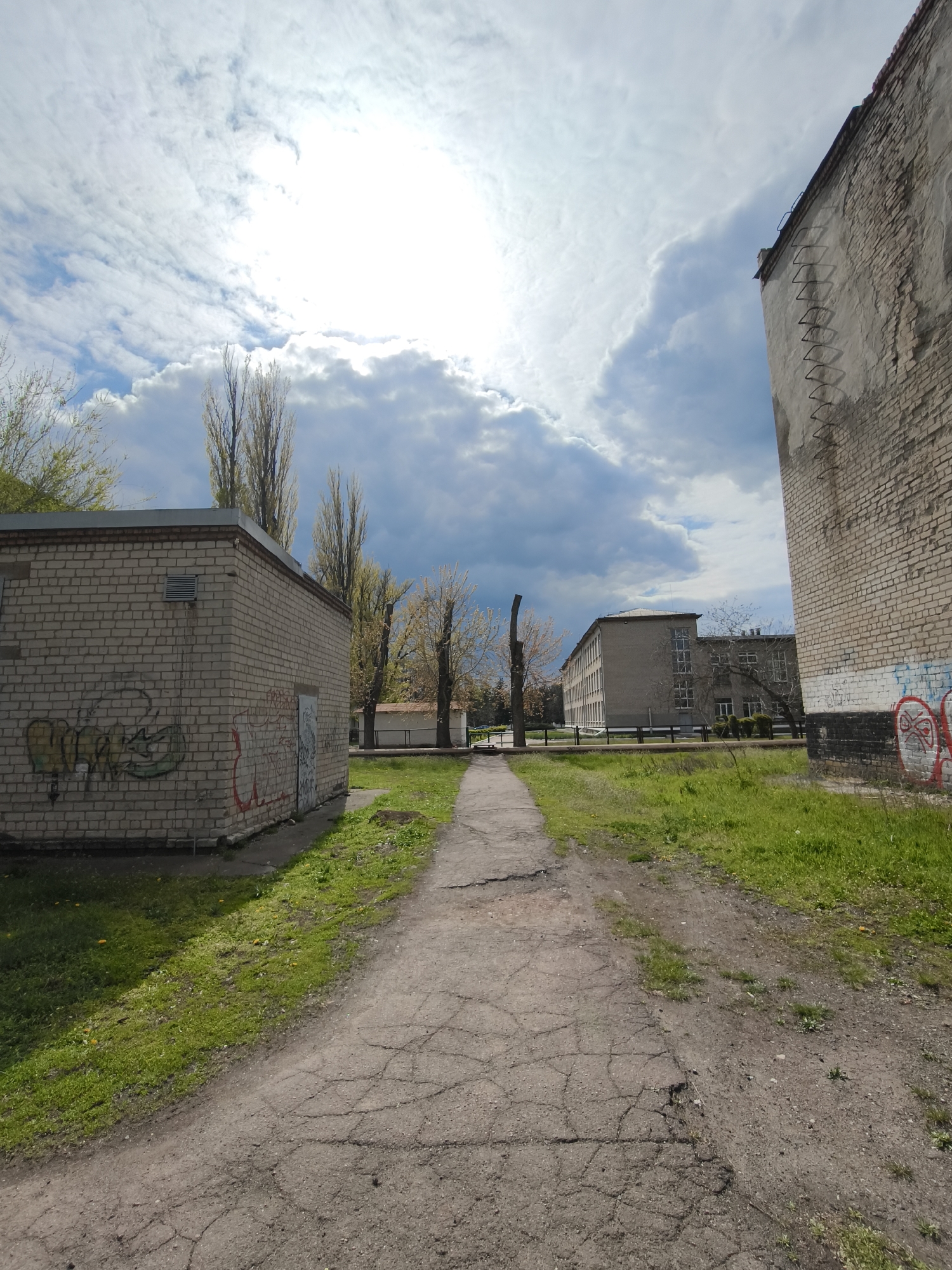 This media file captures an outdoor scene in Kryvyi Rih, Ukraine, featuring a worn asphalt pathway leading into the distance. On the immediate left, a small, single-story brick building is visible, heavily marked with green and yellow graffiti, including the stylized word "GRAFFITI." An air vent is visible on its side. To the right, a taller, partially visible brick building also bears red graffiti at its base, featuring a stylized circular design that resembles a face. A metal ladder is affixed to the upper section of this building.
The path is bordered by patches of green grass, dotted with small yellow flowers. In the mid-ground, tall, slender trees, possibly poplars, line the path, some still bare while others show early spring foliage. Beyond these trees, a larger, multi-story brick building with many windows can be seen in the background, suggesting an institutional or educational facility.
The sky is partially cloudy, with bright sunlight breaking through white and grey clouds, illuminating parts of the scene. No people are visible, and there is no specific activity taking place, lending the scene a quiet, somewhat desolate atmosphere. The cracked pavement and extensive graffiti suggest an aged or industrial urban setting. The time of day is daytime, indicated by the bright sun.