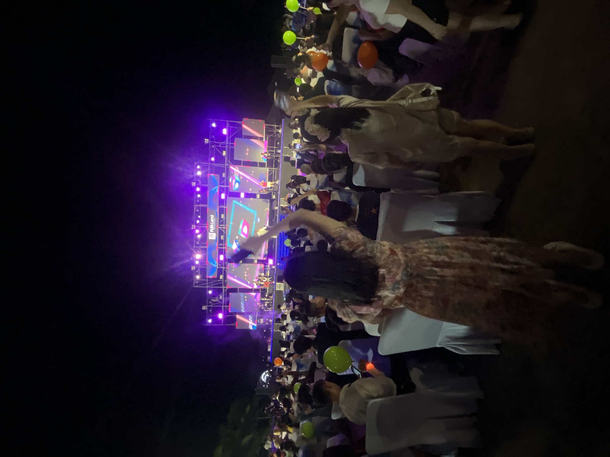 The image depicts a nighttime outdoor event in Da Nang, Vietnam, featuring a live music performance on a brightly lit stage. A large crowd is gathered, seated and standing, to watch the performers. Several people are holding up their phones, presumably to record or photograph the event. Green and orange balloons are visible amongst the audience, adding to the festive atmosphere. The stage is adorned with purple and blue lights, and large screens display colorful graphics. The overall scene suggests a lively and engaging public gathering.