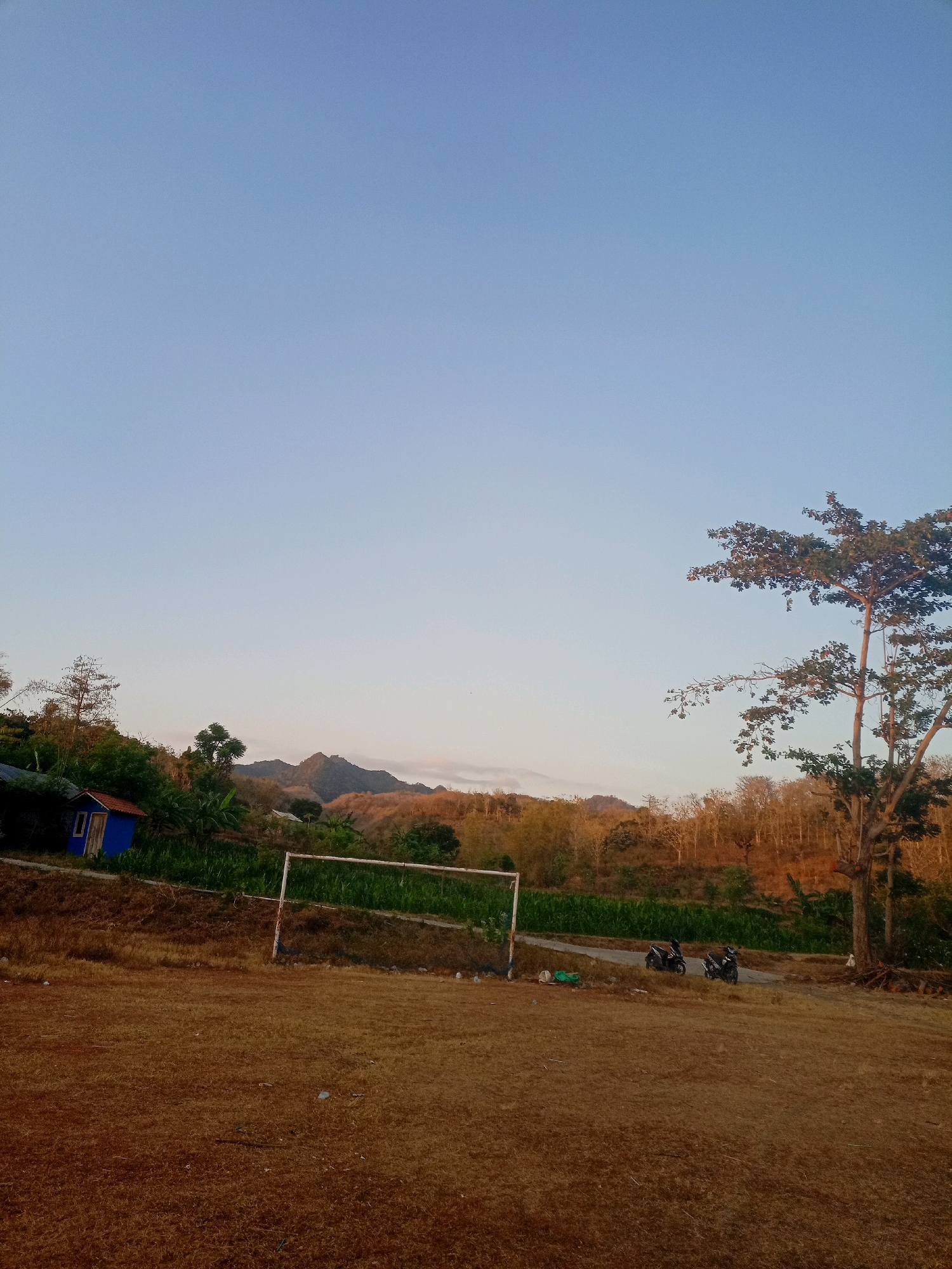 The image depicts an outdoor scene, likely a rural area or outskirts of a town, with a clear sky and soft lighting suggestive of late afternoon or early morning. In the foreground, a dry, brown field dominates, with some scattered debris. A white soccer goalpost stands on the field's edge.
Behind the field, a gentle slope is covered in vegetation, with a small blue building with a red roof situated on the left side. Further back, a road curves through a landscape dotted with trees and agricultural fields. In the middle ground, two motorcycles are parked on the roadside, along with some green bags and other small objects.
The background features rolling hills and mountains, some of which appear dry and sparsely vegetated, while others are covered in dense foliage. A large tree with a broad canopy is prominently positioned on the right side of the frame.
The location is identified as being in the city of Manggar, Indonesia. The overall impression is of a serene, natural environment at the edge of human habitation.