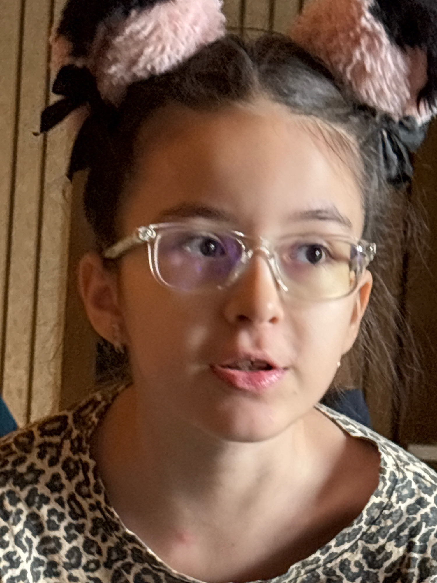 The image shows a close-up portrait of a young girl with brown hair styled in pigtails with fluffy pink and black cat ears attached. She is wearing clear-rimmed glasses and a leopard-print shirt. Her mouth is slightly open as if she is speaking. The background consists of vertical wooden planks. There is no information in the image to indicate the location in the city of Vagator, India.