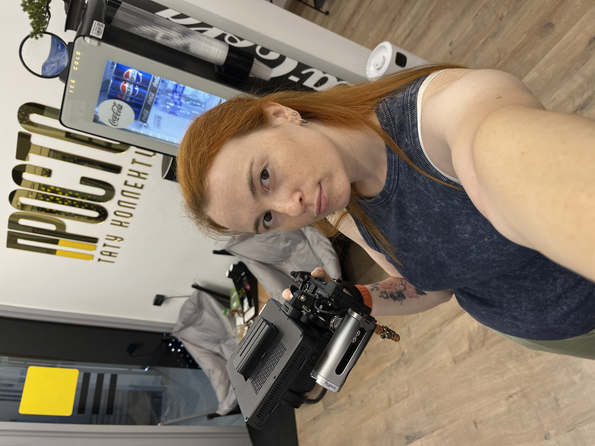 The image shows a young woman with red hair taking a selfie in what appears to be a business establishment. She is holding a professional camera rig with a monitor and a handle. In the background, there is a beverage vending machine with Coca-Cola and Pepsi products, and a sign with golden lettering that reads "ПРОСТО ТАТУ КОЛЛЕКЦИЯ" (Simply Tattoo Collection). There are also chairs and other miscellaneous items in the background, suggesting it could be a waiting area or a break room. The floor is made of light-colored wood. Based on the Russian text on the sign and the context of the establishment, the location is likely a tattoo parlor. However, without further information, it is not possible to determine the exact location within the city of Chaikovsky, Russia.