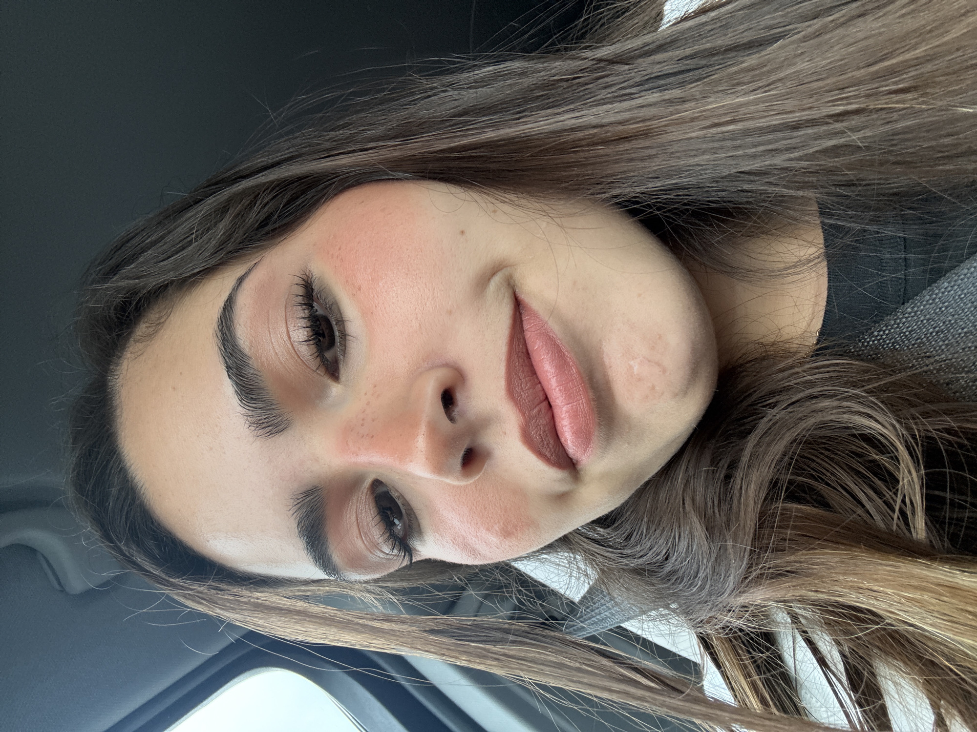 A close-up, angled portrait of a young woman with long brown hair. She is wearing makeup, including noticeable blush and lipstick. The background indicates she is likely in a car, suggested by the car interior elements such as the dashboard and seatbelt visible. No specific details suggest New Braunfels, United States of America. The scene focuses primarily on the subject's face and immediate surroundings within the vehicle.