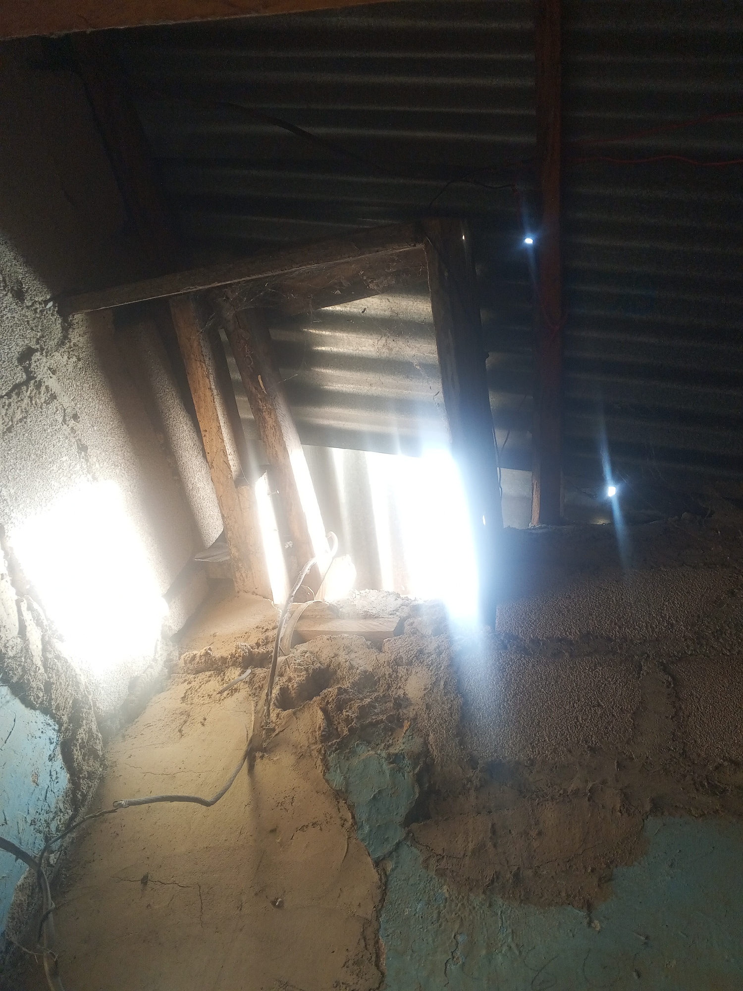 The interior of a building in Gwong, Nigeria is exposed to bright sunlight piercing through openings in the corrugated metal roof and gaps in the rough wall structure. The walls are constructed with a mix of materials including mud, plaster, and wood. One wooden beam is visible, suggesting the roof structure. An electrical cord runs along the wall. The plaster shows signs of age with cracks and peeling.