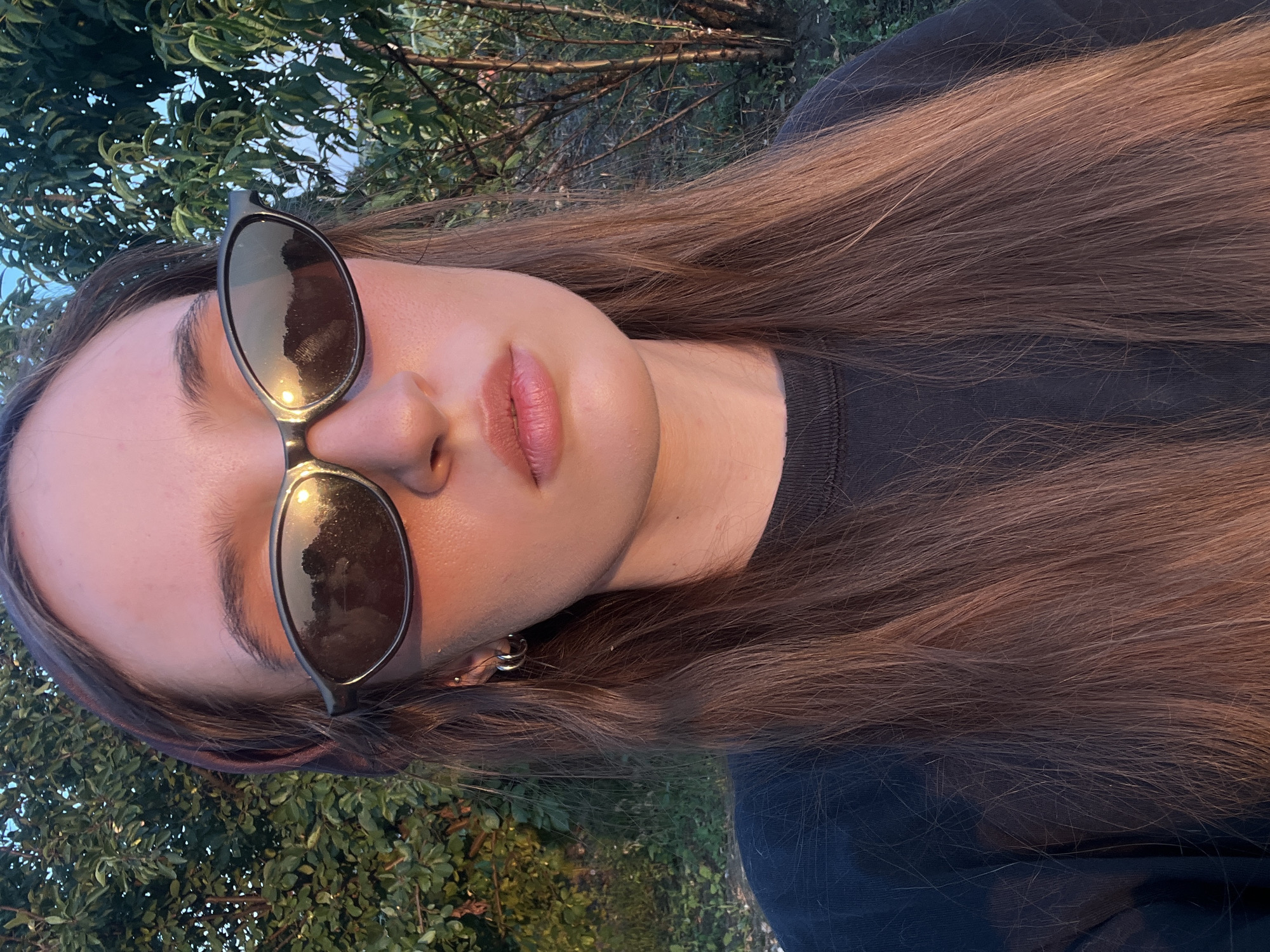 A young woman, viewed from the shoulders up, is outdoors during golden hour, with trees and foliage in the background. She has long, brown hair, a dark sweatshirt, and is wearing oval-shaped sunglasses that reflect the setting sun. Her skin is illuminated by the warm light, creating an orange glow. She has light-colored hoop earrings and a stud earring in her left ear.