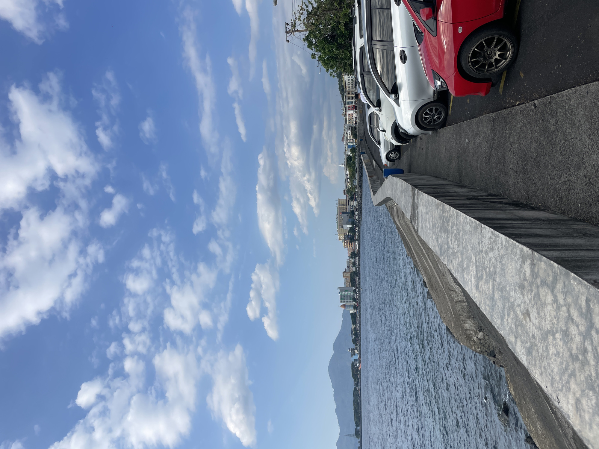 A waterfront promenade lined with parked cars overlooks the ocean and a distant cityscape. The promenade, constructed of concrete, curves gently along the water's edge. Several cars, including a red one in the foreground and a row of silver and white vehicles further down, are parked on the asphalt path adjacent to the promenade. The ocean is calm with small waves lapping against the shore. In the distance, a city skyline with buildings and a large mountain range is visible under a bright blue sky dotted with white clouds. This scene is located in Manado, Indonesia, likely along the boulevard or a similar coastal area.
