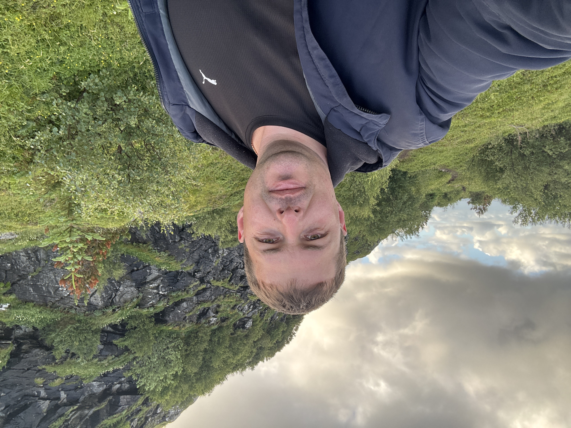 An outdoor, waist-up, front-facing selfie shows a man with light brown hair and a fair complexion. He is wearing a dark blue hooded jacket over a black t-shirt with a white Puma logo. The man is looking directly at the camera with a neutral expression. Behind him, a rocky mountainside covered in green foliage and small trees slopes upwards. The sky is overcast with scattered clouds, with patches of blue visible. The foreground features a grassy slope with scattered small yellow flowers. The setting appears to be the natural landscape of Lovund, Norway, characterized by its mountainous terrain and lush greenery.