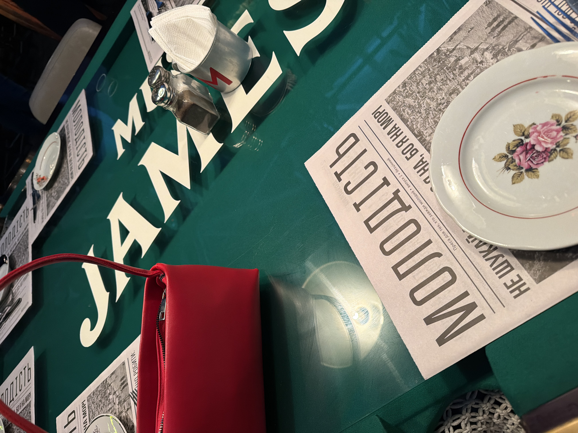 The image shows a close-up of a green table with white lettering that appears to spell out "JAM". A red handbag is placed on the left side of the table, with its zipper visible. Next to the handbag are two small glass shakers, likely for salt and pepper, and a metal cup filled with white napkins. In the foreground, a newspaper is laid out, with the prominent headline in large Cyrillic letters reading "МОЛОДІСТЬ" (Molodist, meaning youth). Below this, smaller text indicates "Преса для тих, хто завжди молодий • випуск 2 • безцінний" (Press for those who are always young • issue 2 • priceless). Further down, a phrase reads "НЕ ШУКАЙ НА, БО Я НА МОРІ" (Don't look for her, because I am at the sea). A small, white decorative plate with a floral pattern is placed on top of the newspaper. The overall impression is of a restaurant or cafe setting in Kyiv, Ukraine, with a modern aesthetic suggested by the table's design and the newspaper's layout.