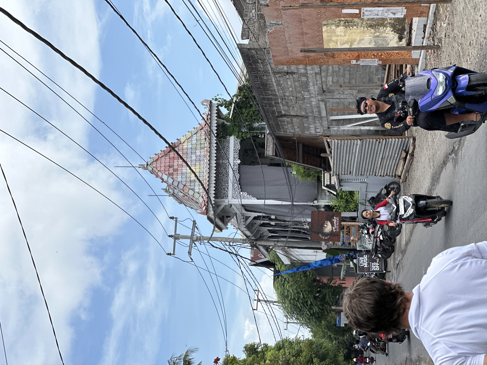 The image captures a street scene in Nusa Penida, Indonesia. Overhead, a web of electrical wires crisscrosses the view. On the left, a building with a distinctive multi-colored tiled roof is visible, alongside a faded exterior and decorative white trim. Various signs and advertisements, including one for "Mambo Dive Resort," are displayed.
People are present on the street, riding motorbikes. One individual is wearing a black shirt and sunglasses, riding a purple motorbike. Another person is visible on a black motorbike with a white and red shirt. A pedestrian's back is visible. Several parked motorbikes line the side of the road. Lush green trees fill the lower part of the image. A clear blue sky with scattered clouds forms the background.