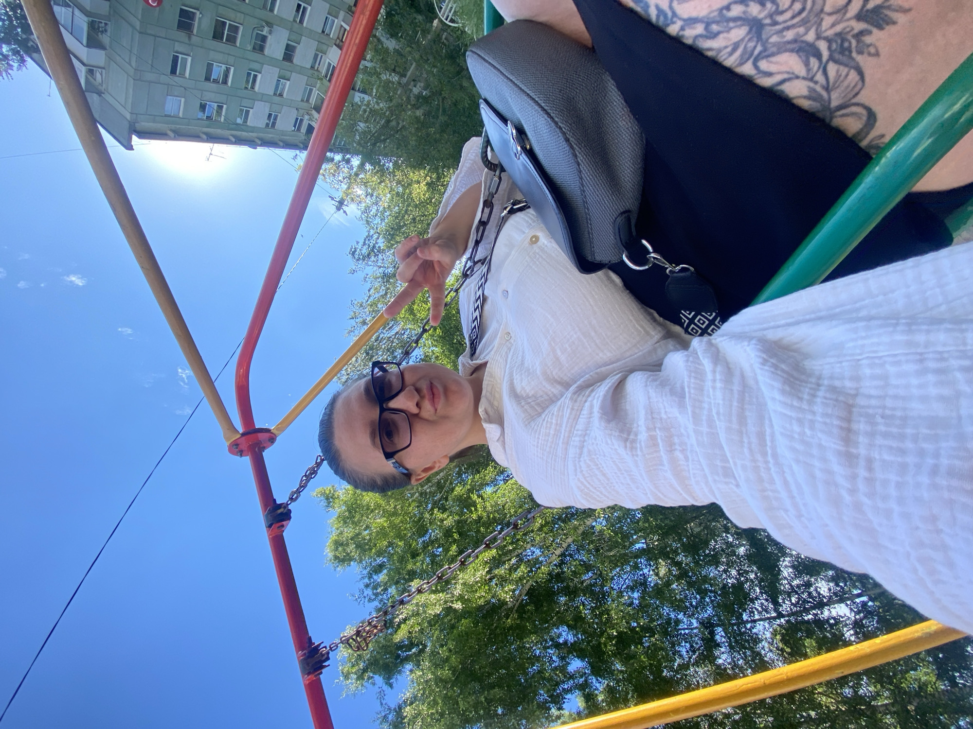 A woman with glasses and a white shirt is taking a selfie outdoors on a sunny day. She is wearing a black crossbody bag. Her left hand is raised, making a peace sign. Behind her, a building and trees are visible under a clear blue sky. The image is taken from a low angle, possibly on a playground or swing set, as indicated by the colorful metal bars and chains in the foreground. The sun is bright, casting a strong glare. There is no visible text in the image.
