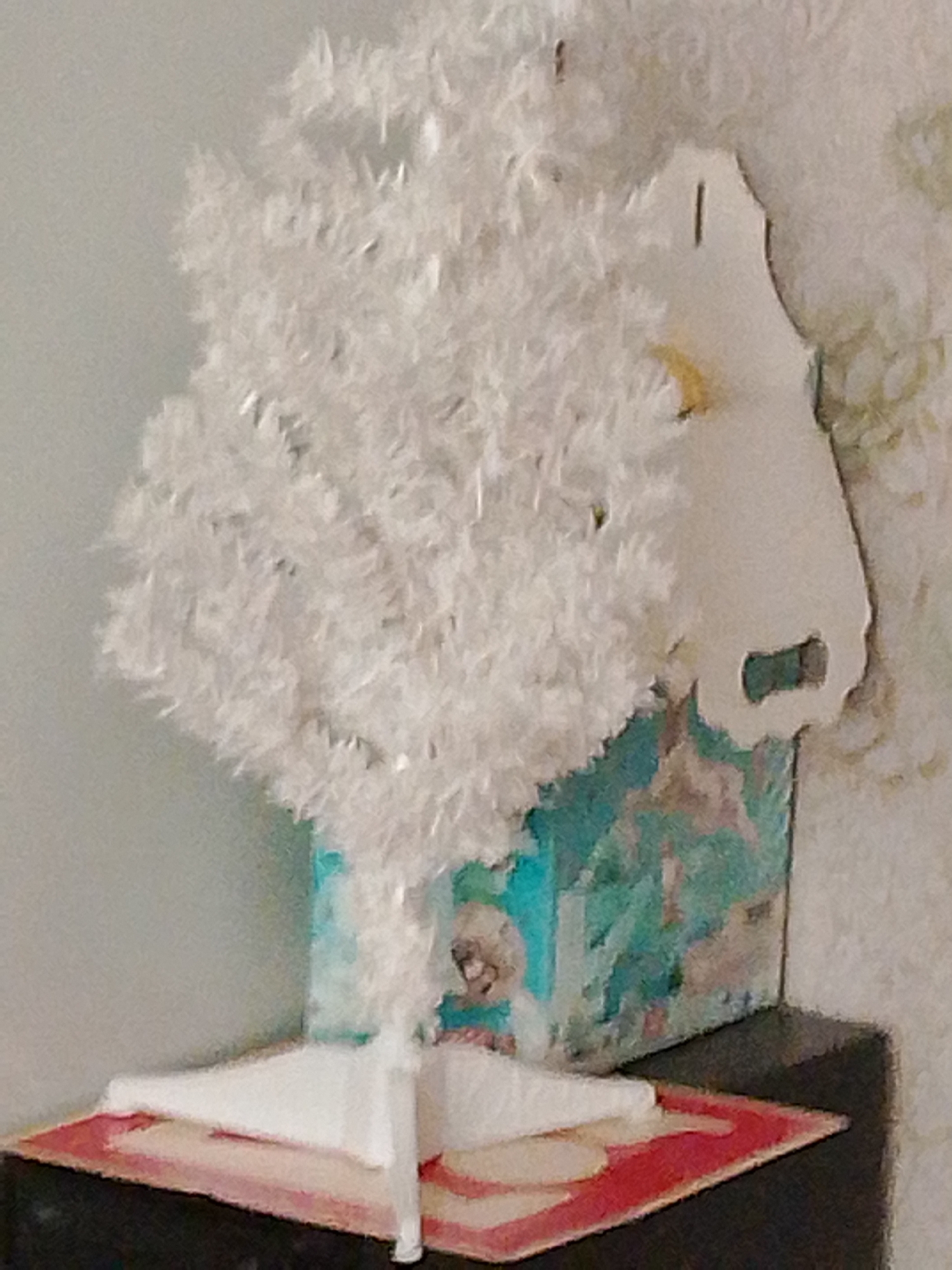 The image shows a small, white, artificial Christmas tree situated on a black surface, possibly a table or shelf. The tree has a fluffy, tinsel-like texture. It is supported by a white stand and placed inside a decorative box or container with a turquoise and patterned exterior, featuring what appears to be animal imagery. A light-colored, cut-out cardboard decoration, possibly a festive ornament, is attached to the right side of the tree's base or the background. The wall behind the tree is textured and light-colored. The scene is lit from the front, creating a shadow on the wall to the right. No people are visible in the image. The overall setting appears to be an indoor domestic environment.