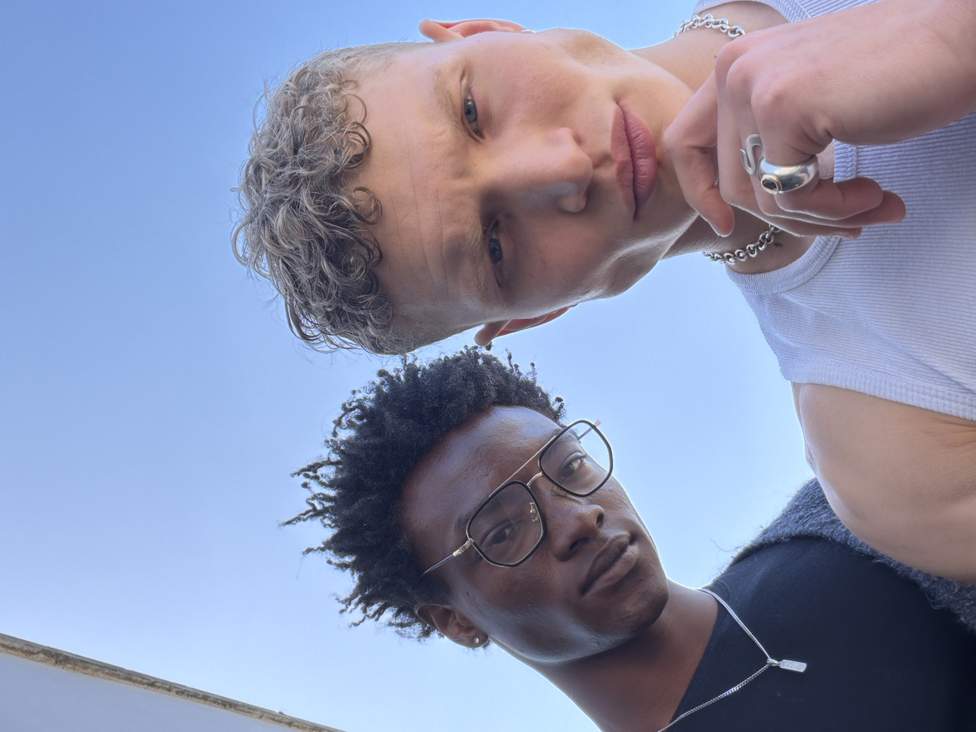 The image features two young men captured from a low-angle perspective against a clear, bright blue sky, indicating daytime and good weather. Both subjects are looking down towards the camera with neutral to slightly contemplative expressions.
The man positioned in the upper frame has fair skin, light-colored curly hair, and blue eyes. He is wearing a white ribbed sleeveless top and accessorized with a silver chain necklace, a silver bracelet on his left wrist, and a prominent silver ring with a dark stone on his right index finger.
The second man, positioned in the lower part of the frame, has a darker skin tone and dark, curly afro-textured hair. He is wearing square-framed glasses with a black and gold design, a black t-shirt, and a silver chain necklace with a small rectangular pendant. A small silver stud earring is visible in his left ear.
A minimal, light-colored architectural edge, possibly a building or wall, is visible at the very bottom left. The overall scene suggests a casual outdoor setting. There is no visible text in the image. The location context is London, United Kingdom.