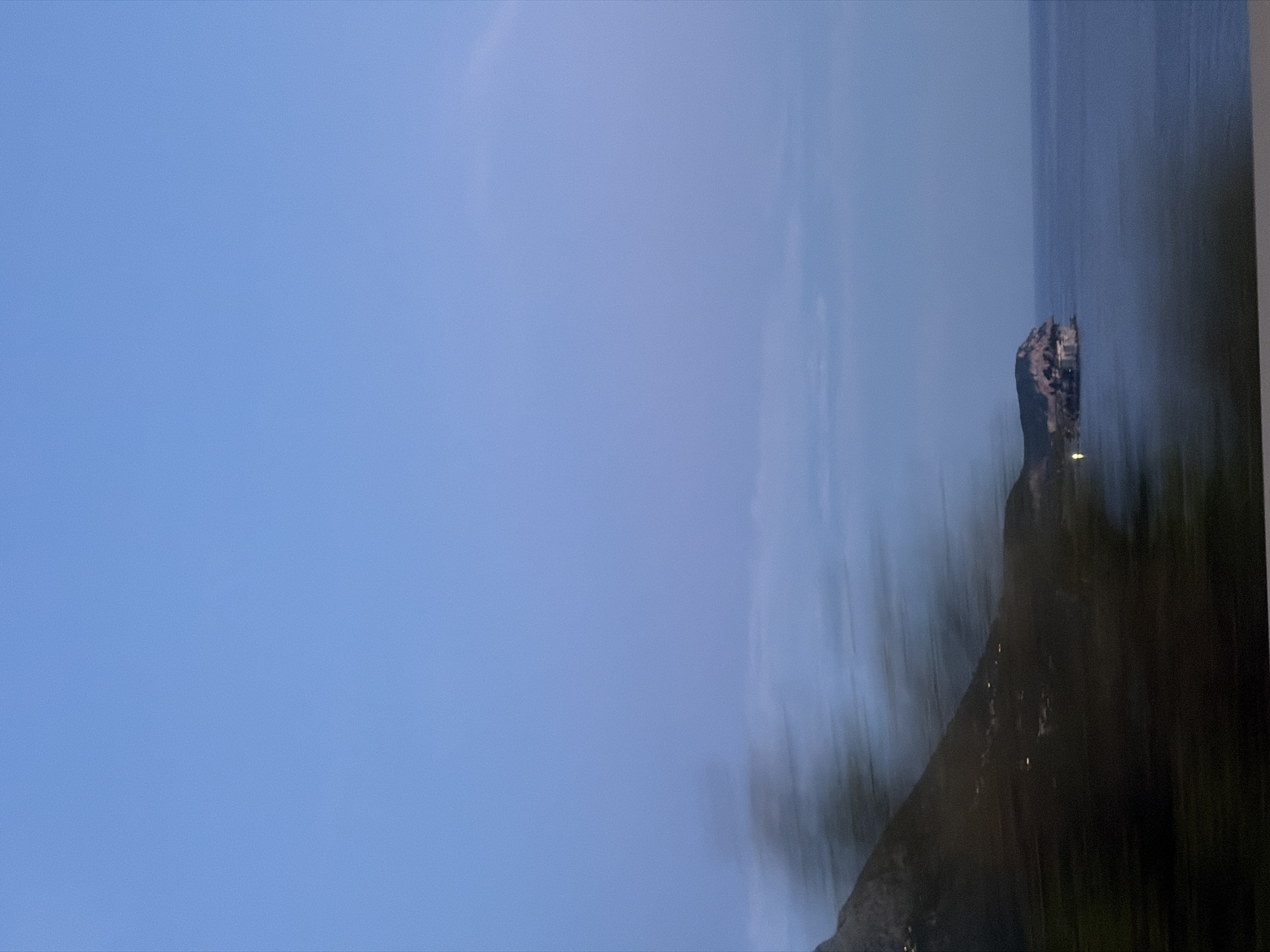 The image depicts a coastal landscape at dusk. The sky is a clear, pale blue, fading into a lighter hue near the horizon. A vast expanse of calm, dark blue water occupies the lower right portion of the frame. On the left, a steep, dark mountainside rises, its lower slopes blurred with motion streaks, suggesting movement of the camera or a fast-moving vehicle. Scattered lights are visible along the base of the mountain, indicating a settlement. To the right of the mountain, a rocky peninsula juts out into the sea, with structures visible on its summit. This geological formation is likely the location of Rafailovići, a settlement known for its picturesque coastal setting in Montenegro. The overall impression is one of a serene, twilight scene by the Adriatic Sea, with the dynamic blur on the mountain adding a sense of travel or speed.