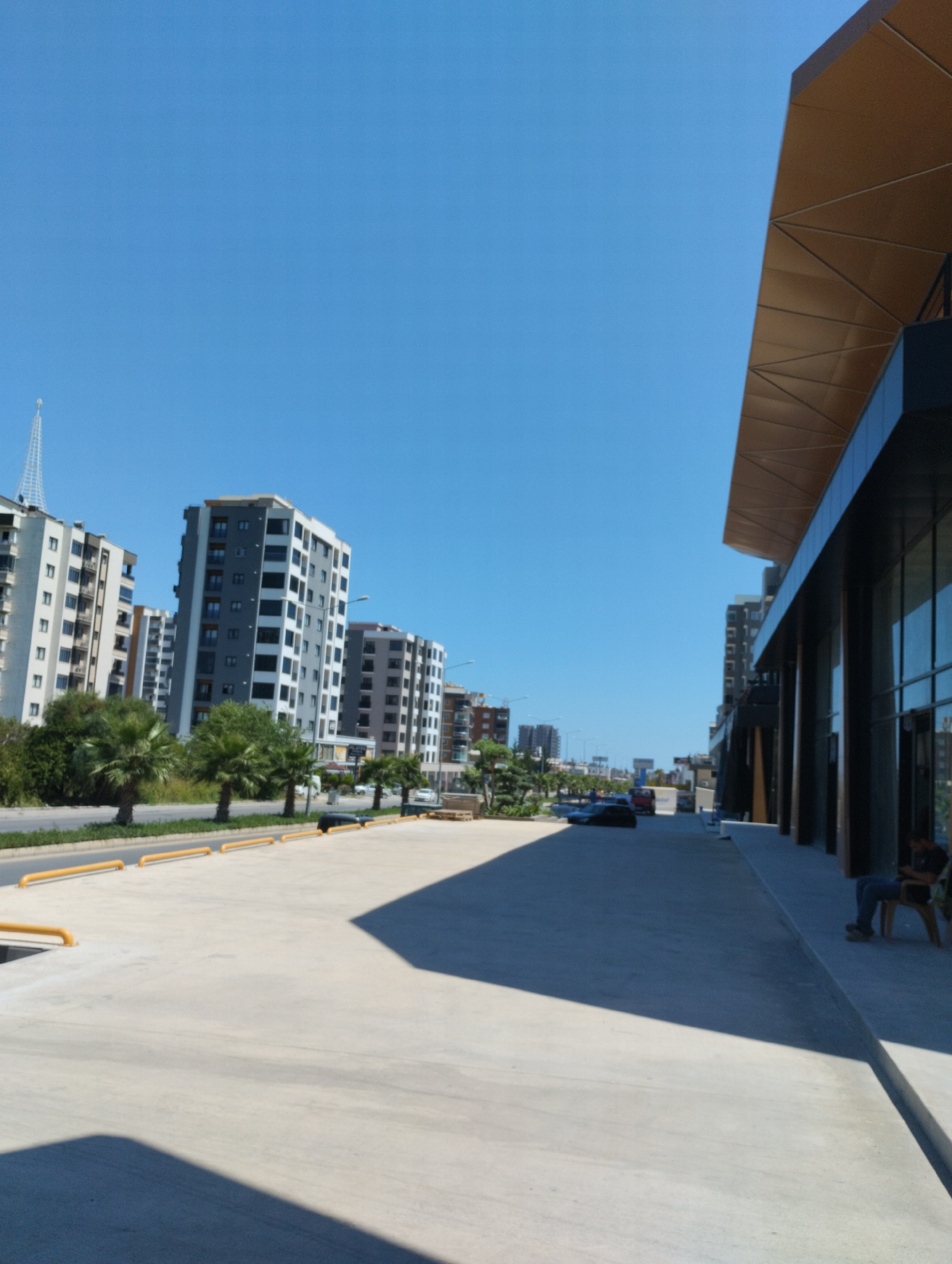 The image depicts an outdoor scene in Toroslar, Turkey, on a clear, sunny day. In the foreground, a vast, light-colored concrete expanse, possibly a parking lot or plaza, dominates the lower portion of the frame, with yellow bollards lining the edge. To the right, a modern building with dark columns and a large glass facade is visible, featuring a striking geometric canopy overhead. A person is seated on a chair on the sidewalk in front of this building.
Further back, a street runs horizontally across the midground, lined with lush green trees and palm trees. Beyond the street, a row of multi-story apartment buildings stands, varying in architectural style and color, with many featuring balconies and gray and white facades. The sky above is a brilliant, cloudless blue. In the distance, more buildings and structures are visible, suggesting a developed urban environment. The overall impression is one of modern construction and urban living.