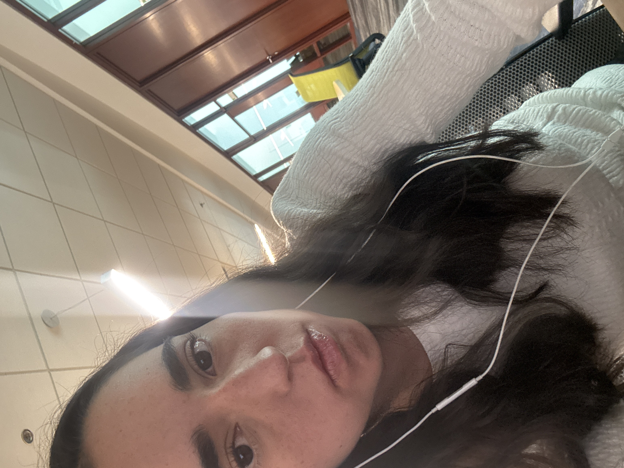An adult female is depicted in a close-up, angled self-portrait within an indoor commercial or office environment. She has long, dark brown hair, dark eyes, and a neutral expression, looking directly into the camera. She is wearing a white, textured long-sleeve top and white wired earbuds, with the cable visible across her upper chest. The background features a light-colored drop ceiling with grid tiles, incorporating two visible linear fluorescent light fixtures, one of which creates a strong lens flare, and several recessed circular lights. Beyond the subject, partitioned walls with brown frames and multiple clear glass panels are visible, suggesting an office or meeting room. Through these glass panels, a bright yellow office chair and additional interior lighting are discernible. Part of a dark office chair with a mesh back is visible in the lower right foreground, indicating the subject's seating. The location is an office or corporate workspace within Unknown, Unknown.